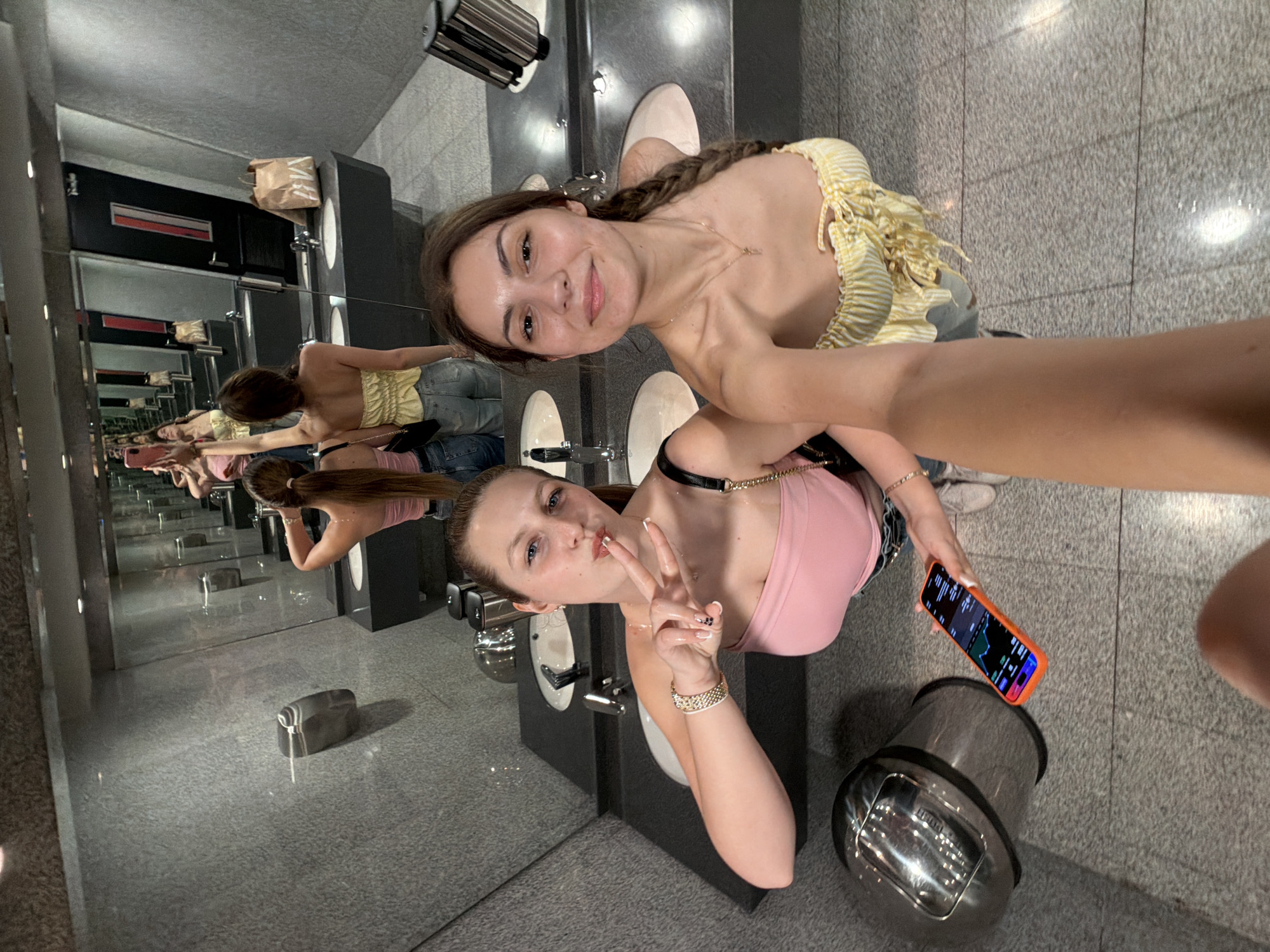 The image captures two young women posing in what appears to be a modern public restroom, taken from an angle that rotates the scene 90 degrees clockwise.
In the foreground, the woman on the left, with long brown braided hair, smiles gently at the camera. She wears a yellow and white striped off-the-shoulder top and light-colored jeans. To her right, the second woman, with light brown hair pulled back, makes a peace sign with her right hand while looking towards the camera with a slight pout. She is wearing a pink tank top, a black choker with a gold chain, and a gold bracelet on her left wrist. In her left hand, she holds an orange-cased smartphone that displays what looks like financial charts or data with green and red lines.
The setting is characterized by a series of dark grey sink counters, multiple faucets, and large mirrors that create an illusion of infinite reflections of the women and the restroom extending into the background. Hand dryers are visible mounted on the walls. A light brown paper bag rests on top of a dark dispenser unit seen in the reflections. The floor is made of grey speckled tiles. A metallic trash can is visible on the floor near the woman in the pink top.
The activity appears to be a casual selfie or photograph session. Both women seem cheerful and engaged with the camera. The bright, artificial lighting suggests an indoor environment, possibly a commercial establishment in Burgas, Bulgaria. No readable text is discernible on any objects other than the graphical data on the smartphone screen.