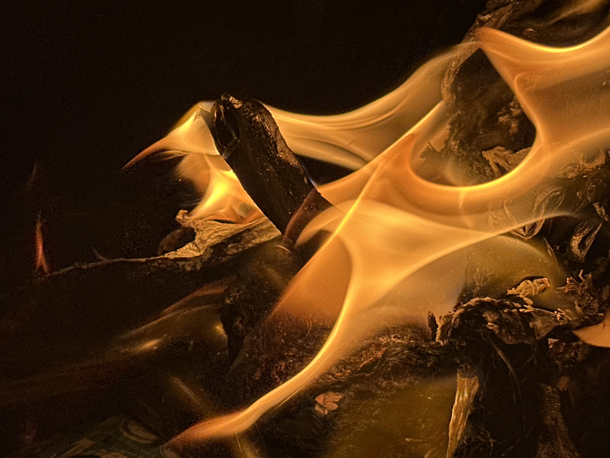 A close-up photograph depicts a dark, low-lit scene dominated by flames. A partially charred, dark object, possibly a piece of wood or similar material, is centrally positioned and engulfed by bright orange and yellow flames. Debris surrounds the charred object, it is located in Bar Gada, Nigeria. No people are visible.