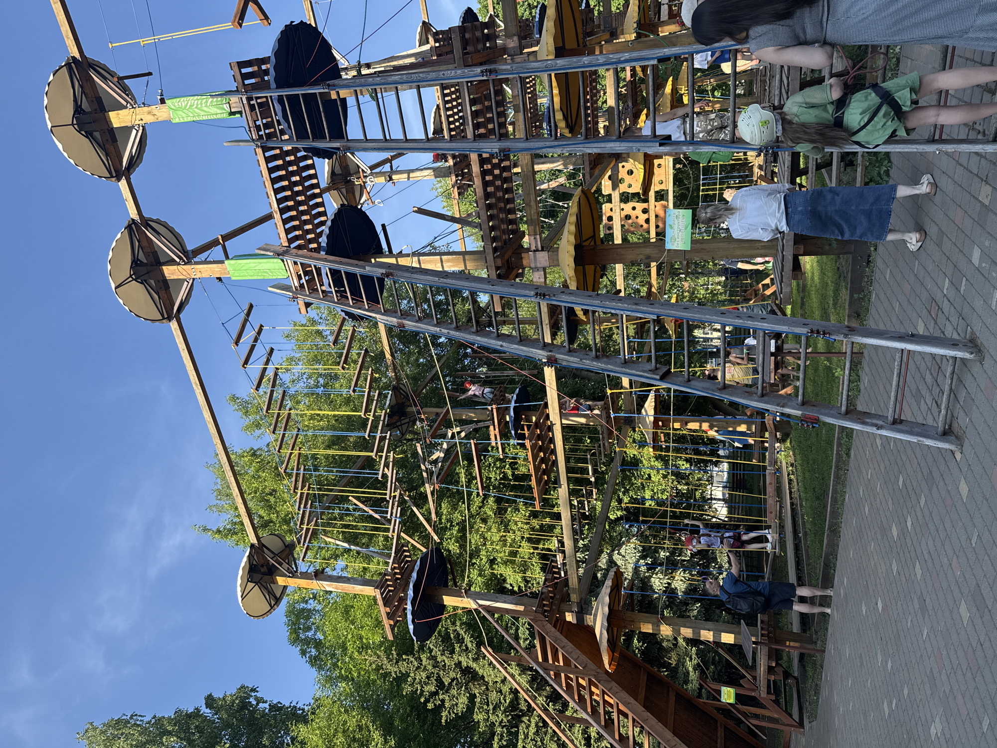 The image depicts a tall, complex rope park structure located in Izhevsk, Russia. The structure is made of wood, metal, and rope, and includes various climbing elements such as ladders, platforms, and rope bridges. Several people are visible on the ground observing the park, while others are navigating the park. The structure is set outdoors with green trees visible behind the play structure and clear blue sky. The ground is paved.
