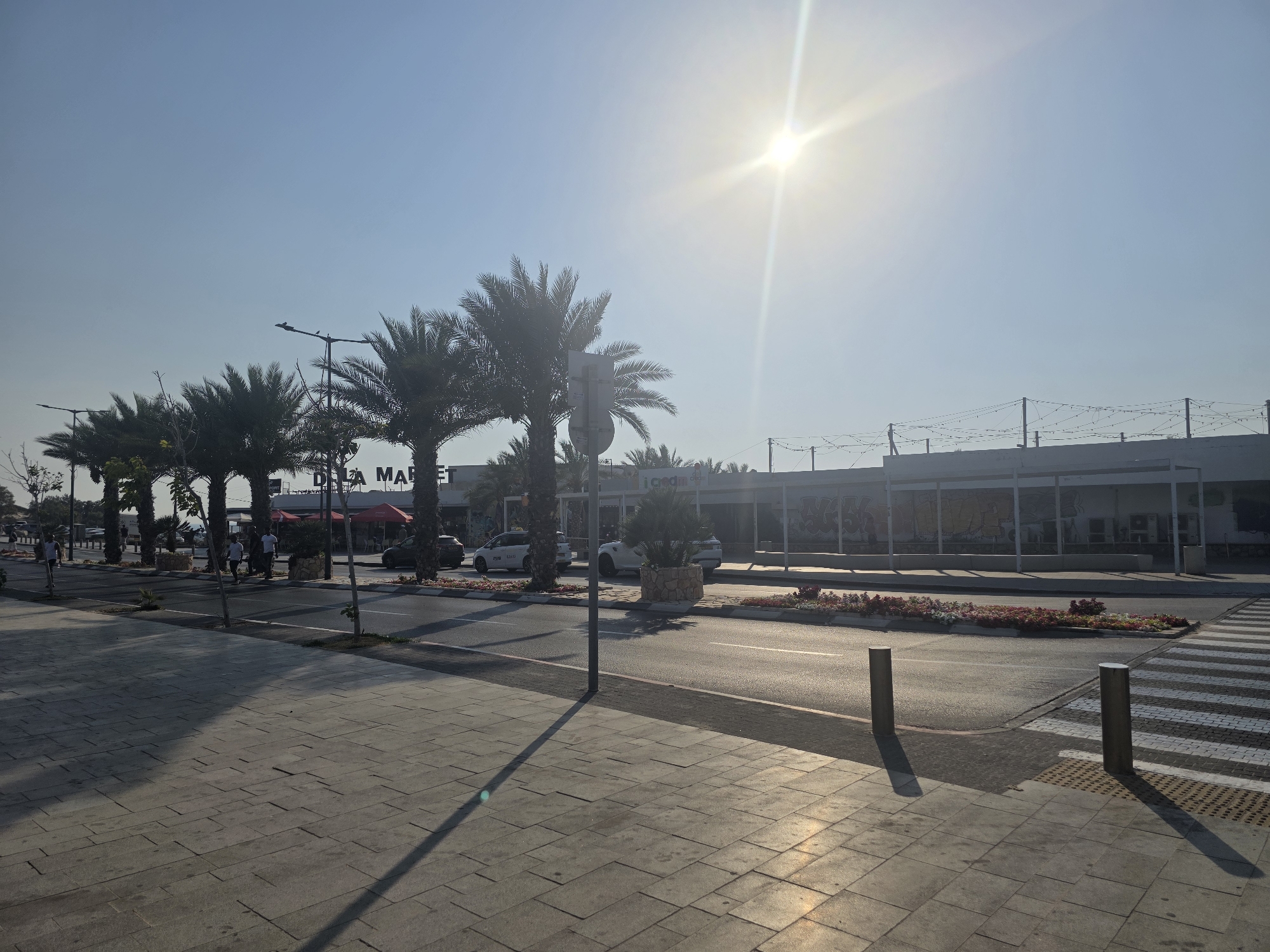 The image depicts a sunny day in Ashkelon, Israel, with a view of a street scene. In the foreground, a wide sidewalk paved with large rectangular stone tiles is visible. The sun's rays cast long shadows across the pavement. A street with a pedestrian crossing is to the right of the sidewalk. Several bollards stand on the edge of the sidewalk, near the crosswalk. Beyond the street, a row of palm trees lines the median strip, with a street light visible above them. In the background, a modern building complex stretches across the scene. The building has a white facade and an open-air walkway with a light string decoration. A sign that appears to read "DILA MARKET" is visible above the entrance to one of the establishments. A few people are walking on the sidewalk and crossing the street. Cars are parked and driving on the street. The sky is clear and bright blue, with the sun prominently positioned in the upper center of the image, creating a lens flare. The overall scene suggests a commercial area on a bright, clear day.