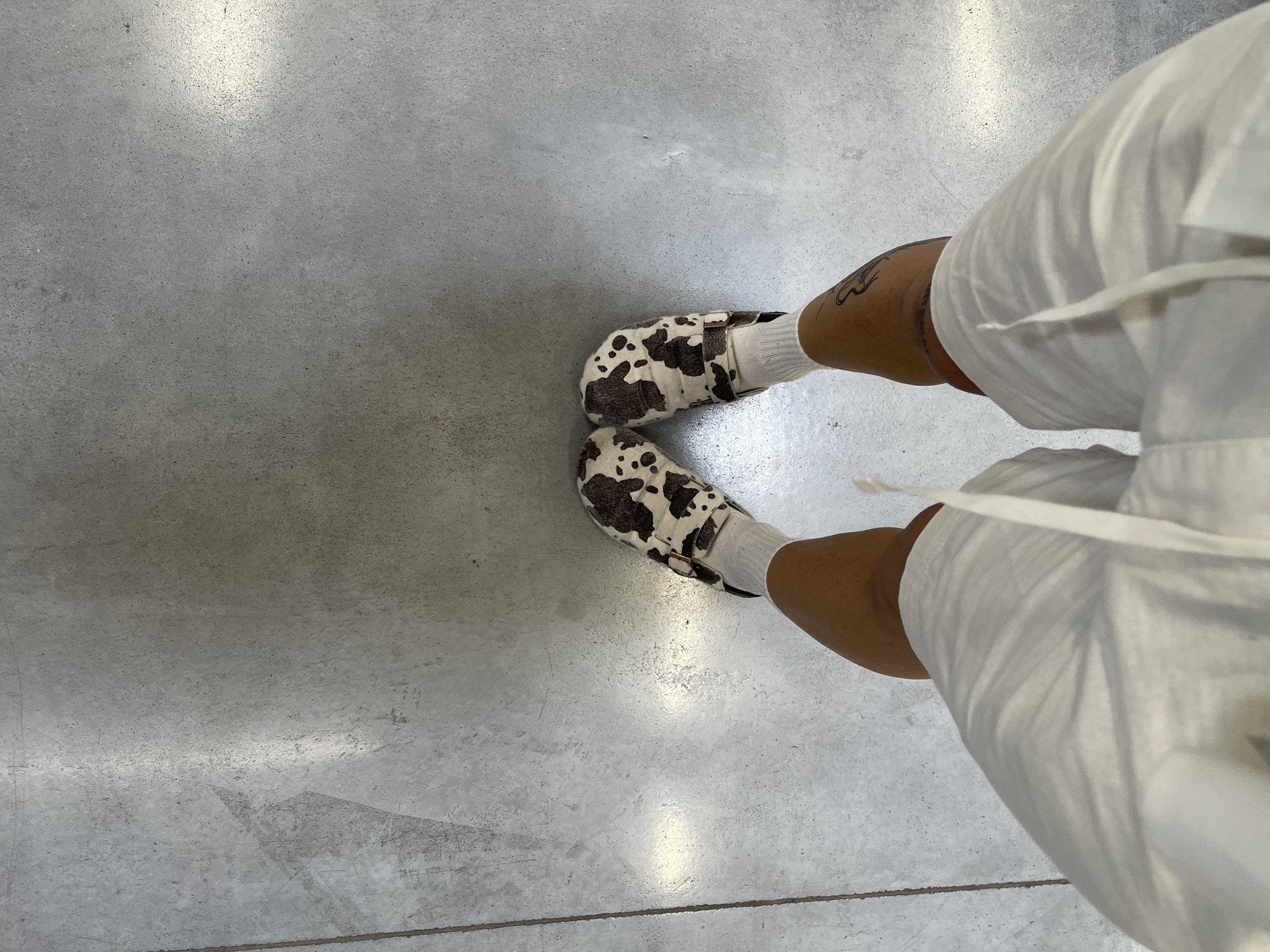 An overhead, medium-level shot captures the lower legs and feet of a person standing on a light grey concrete floor. The person wears white ankle socks and distinctive slip-on shoes with a cow-print pattern, characterized by irregular dark brown or black patches on a cream or white base. Each shoe features a brown strap and buckle detail across the instep. On the right side of the frame, the lower portion of a white garment, possibly shorts or pants with a visible drawstring, is present. The individual's skin tone is medium to dark, and a dark, stylized tattoo is visible on the outer calf of the left leg, just above the ankle. The concrete floor exhibits a fine grain texture with subtle reflections, indicating either an indoor setting or a well-lit covered outdoor area. Minor scuff marks and variations in the concrete's tone are discernible. The image captures a casual moment, emphasizing the person's footwear and lower attire. No text is visible in the image. The location is Su Masu/Elmas, Italy.