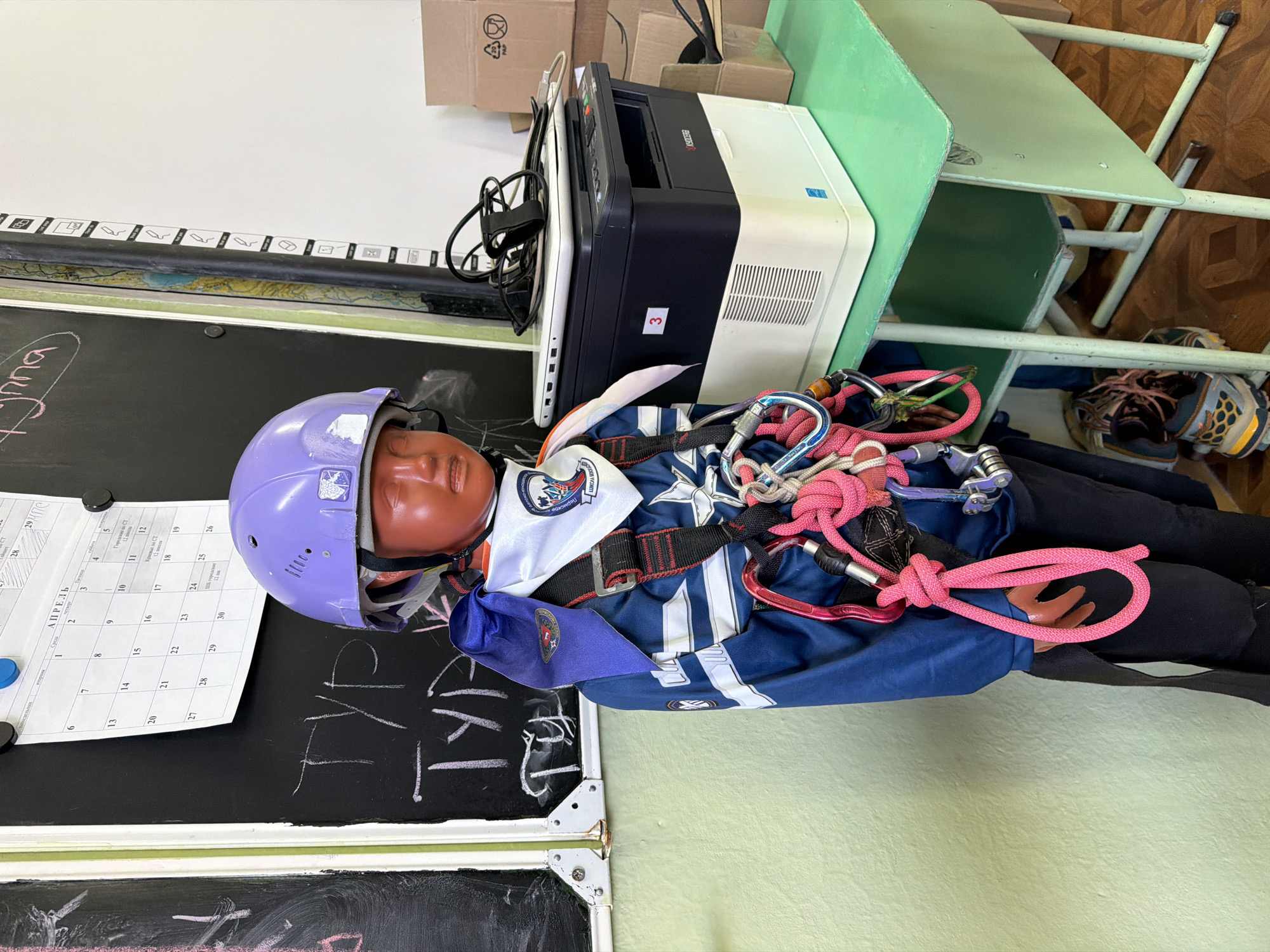 The image shows a training dummy wearing a purple helmet and a blue and white harness, lying on a surface next to a chalkboard. The chalkboard has a calendar attached and writing in chalk. A printer is visible behind the dummy, along with a green desk and a pair of shoes. The location appears to be an indoor classroom-like setting. There is no definitive evidence to confirm the location as the city of Berezniki, Russia.