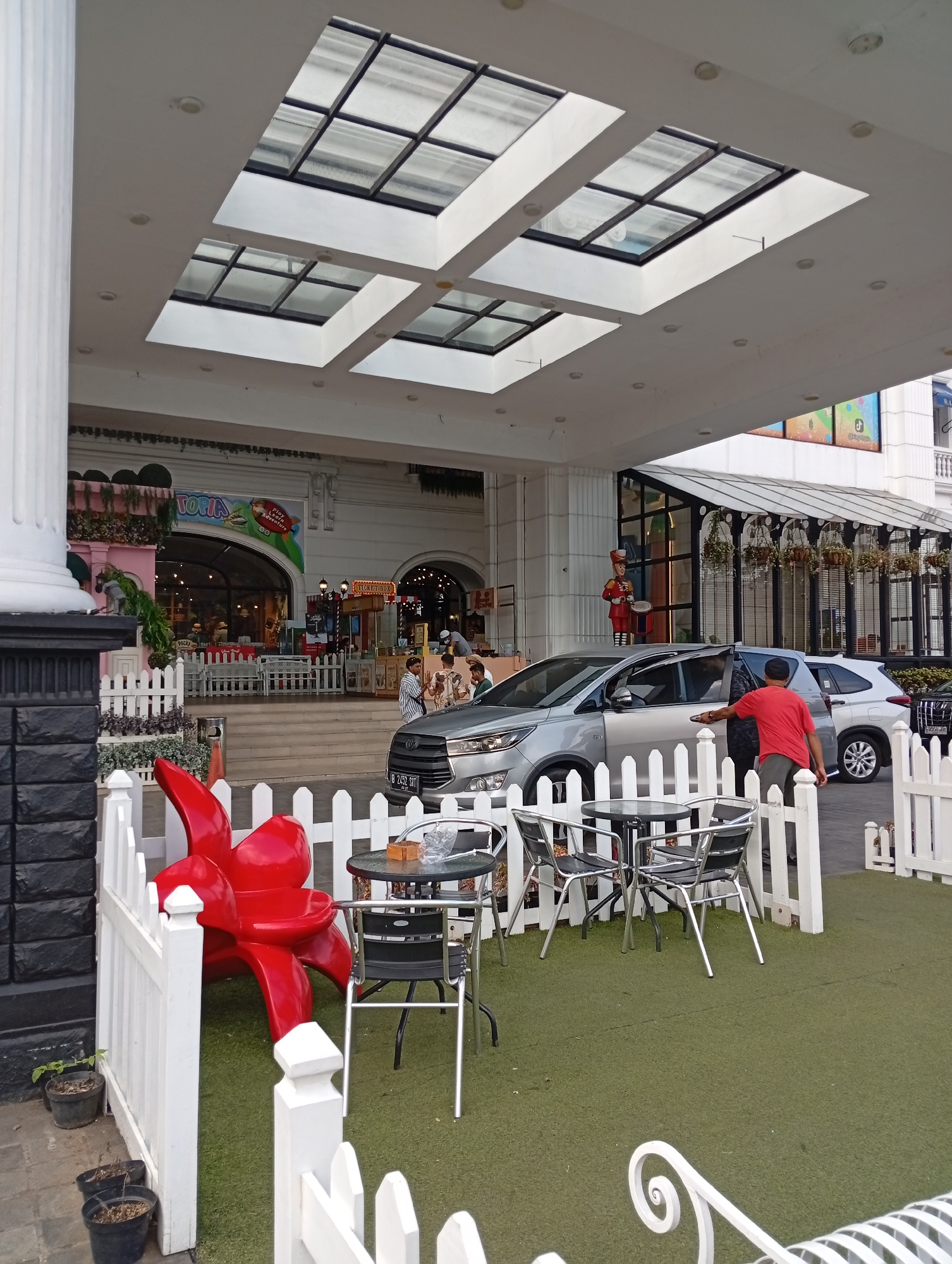 The image captures an outdoor scene in front of a building, likely a commercial establishment, in Cimacan, Indonesia. A white picket fence encloses a section of artificial turf, where a red sculptural flower-like object is prominently displayed. A round black table with four chairs, two of which are black and two are silver, sits on the turf. A man in a red t-shirt and grey pants is seen opening the car door of a silver multi-purpose vehicle (MPV) parked nearby. A white SUV is parked behind the silver MPV. The building behind the fence has a white facade with large glass windows and a white roof with multiple skylights. In the background, some commercial signage is visible, including one with the word "UTOPIA". A few potted plants are placed near the fence and on the walkway. The overall impression is an entrance area with a decorative outdoor seating space.