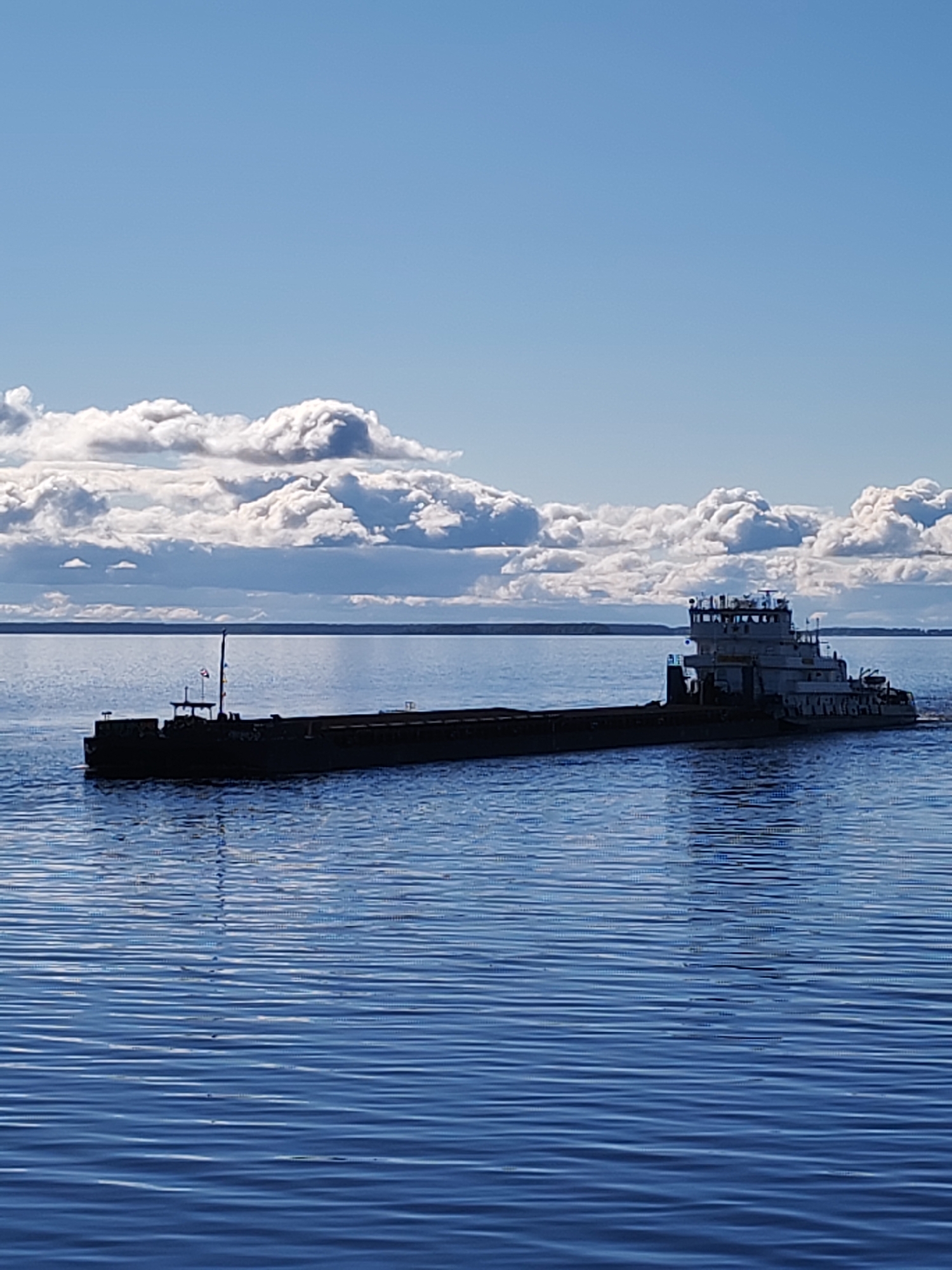 A long, flat-bottomed barge with a tall superstructure located at the rear is traversing a body of water under a partly cloudy sky. The barge is dark, possibly black or a dark shade of gray. The water is a deep blue color, showing distinct wave patterns. The sky is light blue with large, fluffy white clouds clustered together. A strip of land is faintly visible in the distance, just above the waterline. This image shows a scene near Mikhalevo, Russia.
