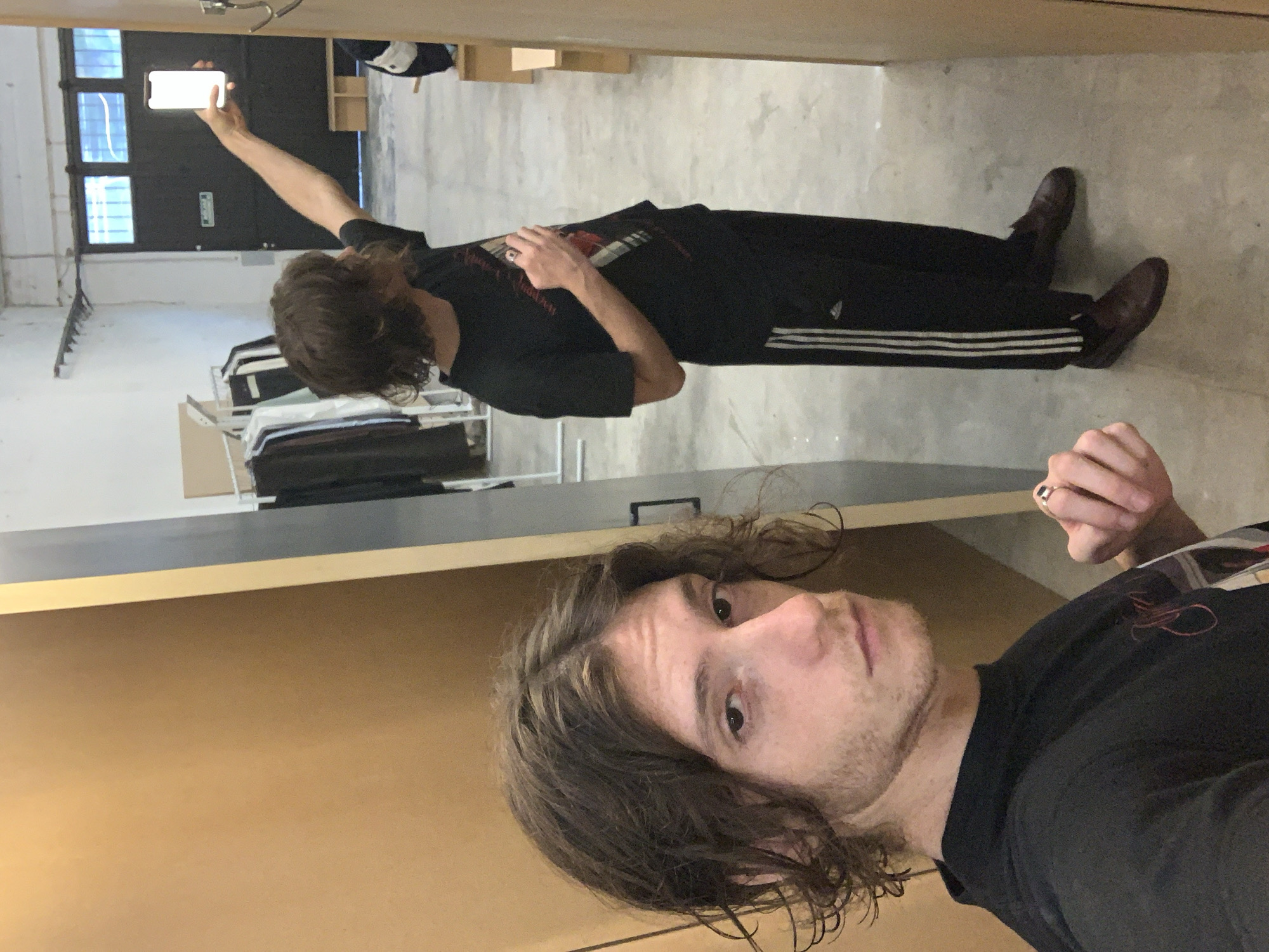 A selfie-style photograph, taken indoors, features two young men. In the foreground, a man with medium-length wavy brown hair and light stubble looks directly at the camera with a neutral expression. He wears a black t-shirt with a red graphic and a ring on his right hand.
Behind him, slightly out of focus, stands another man with similar brown wavy hair. He is dressed in a black t-shirt and black track pants with three distinctive white Adidas stripes running down the side. He wears dark brown shoes. His right arm is extended upwards, holding a brightly illuminated smartphone, as he appears to be capturing an image or video above him, his gaze fixed on the screen.
The setting is an indoor space with a concrete floor and light-colored walls. A prominent light brown wooden partition occupies the immediate foreground. In the background, large dark doors or windows with white frames are visible, along with a shelving unit holding folded clothing. A small, white rectangular sign with illegible green text is affixed to the dark background wall. The scene suggests a utilitarian room, possibly a studio or workshop. The image originates from Buenos Aires, Argentina.