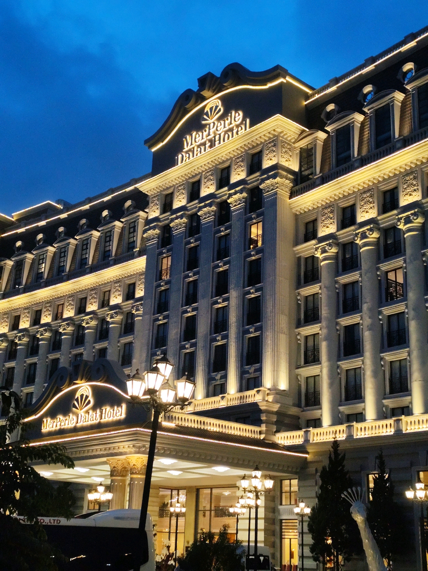 The image shows the MerPerle Dalat Hotel in Da Lat, Vietnam at dusk. The hotel is a large, ornate building with a classical facade, illuminated by warm yellow lights. The architecture features multiple stories with arched windows, decorative columns, and a prominent mansard roof with dormer windows. A sign with the hotel's name, "MerPerle Dalat Hotel," is lit up above the entrance. In front of the hotel, there are several lampposts with multiple lights. A white bus is partially visible in the foreground, along with some dark green trees and a unique, abstract sculpture that appears to be reaching upwards. The sky is a deep blue, indicating the transition from day to night.