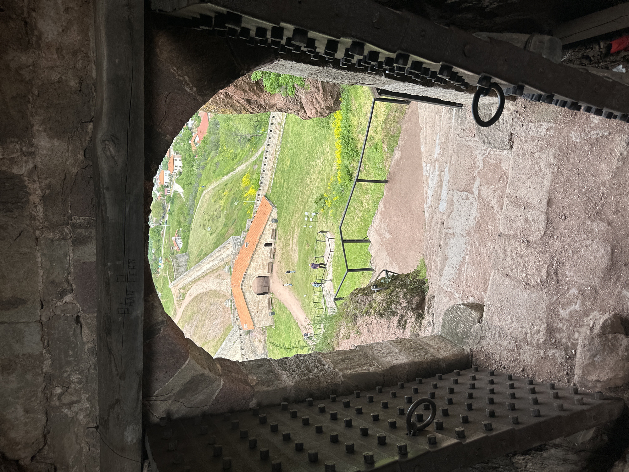 This eye-level, close-up shot, taken from within the Belogradchik Fortress in Belogradchik, Bulgaria, looks outward through a sally port or similar opening. The opening, framed by rough-hewn stone, has a metal grate with studs and ring pulls on the bottom and top, possibly a portcullis-like mechanism. Outside, the view encompasses a section of the fortress grounds, including grass, a brick wall, a pathway, and distant structures. In the distance, there are buildings and greenery, indicating the town or landscape surrounding the fortress. The lighting is somewhat dim inside the fortress compared to the brighter outdoor light. There are no people visible within the fortress framing the view; the focus is solely on the external landscape and architectural elements of the fortress. There are people walking around the fort, visible by the orange wall.