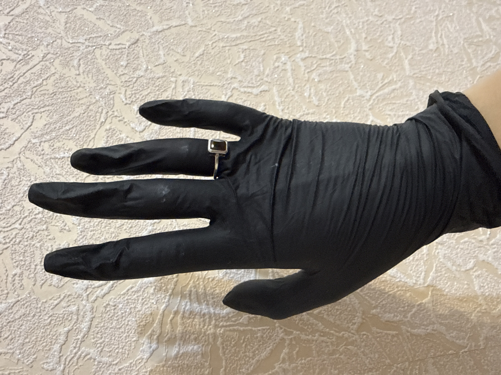 The image shows a close-up of a gloved hand. The hand is wearing a black glove and is positioned in front of a textured, light-colored background. A small, silver ring with a rectangular, dark gemstone is visible on the ring finger. The glove appears to be made of latex or nitrile material, and there are some faint smudges on its surface. The background has a raised, irregular pattern that resembles wallpaper or a textured wall. The lighting suggests an indoor setting, possibly with artificial light, as there are no strong shadows indicating natural sunlight. The image is focused on the hand and the ring, with the background slightly out of focus, emphasizing the jewelry. The overall impression is that of a detail-oriented shot, possibly for product photography or showcasing the ring. No specific time of day, weather, or location cues are evident. There is no visible text in the image.