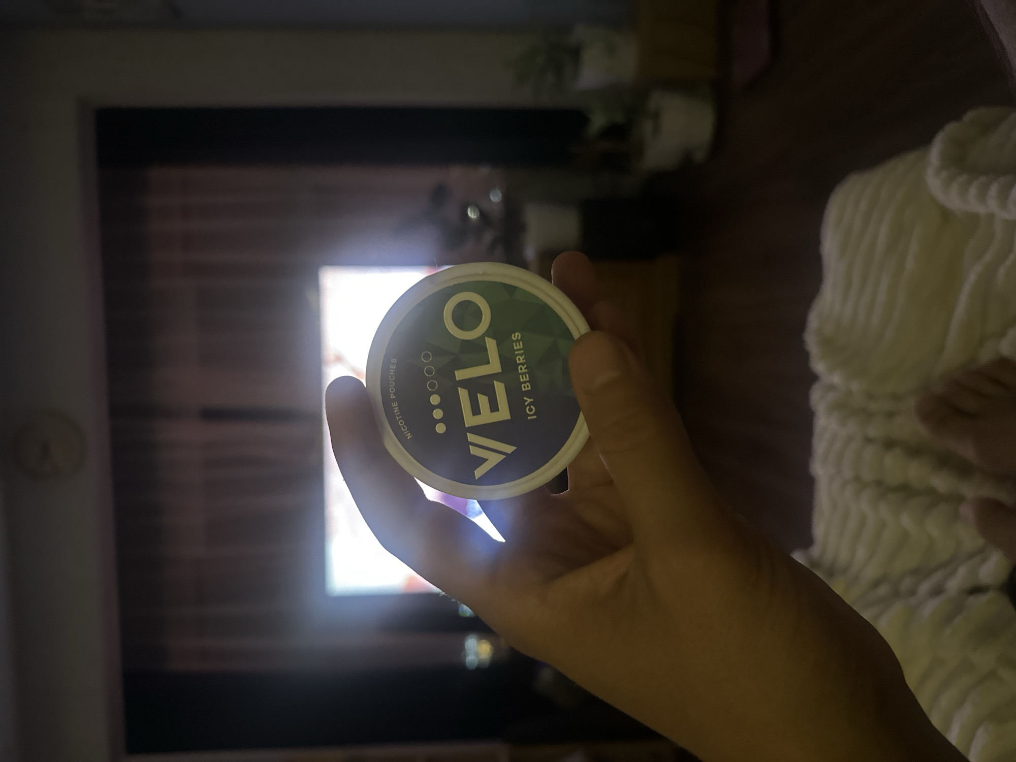 A person's hand is holding a round, dark blue and green container of VELO nicotine pouches. The container has text on it that reads "NICOTINE POUCHES" and "VELO ICY BERRIES". The background is blurred and appears to be a living room with a television screen on and some potted plants. The person's feet are also visible in the foreground, resting on a white, textured blanket. There is no indication of the location being in Nam Nha Trang Ward, Vietnam.