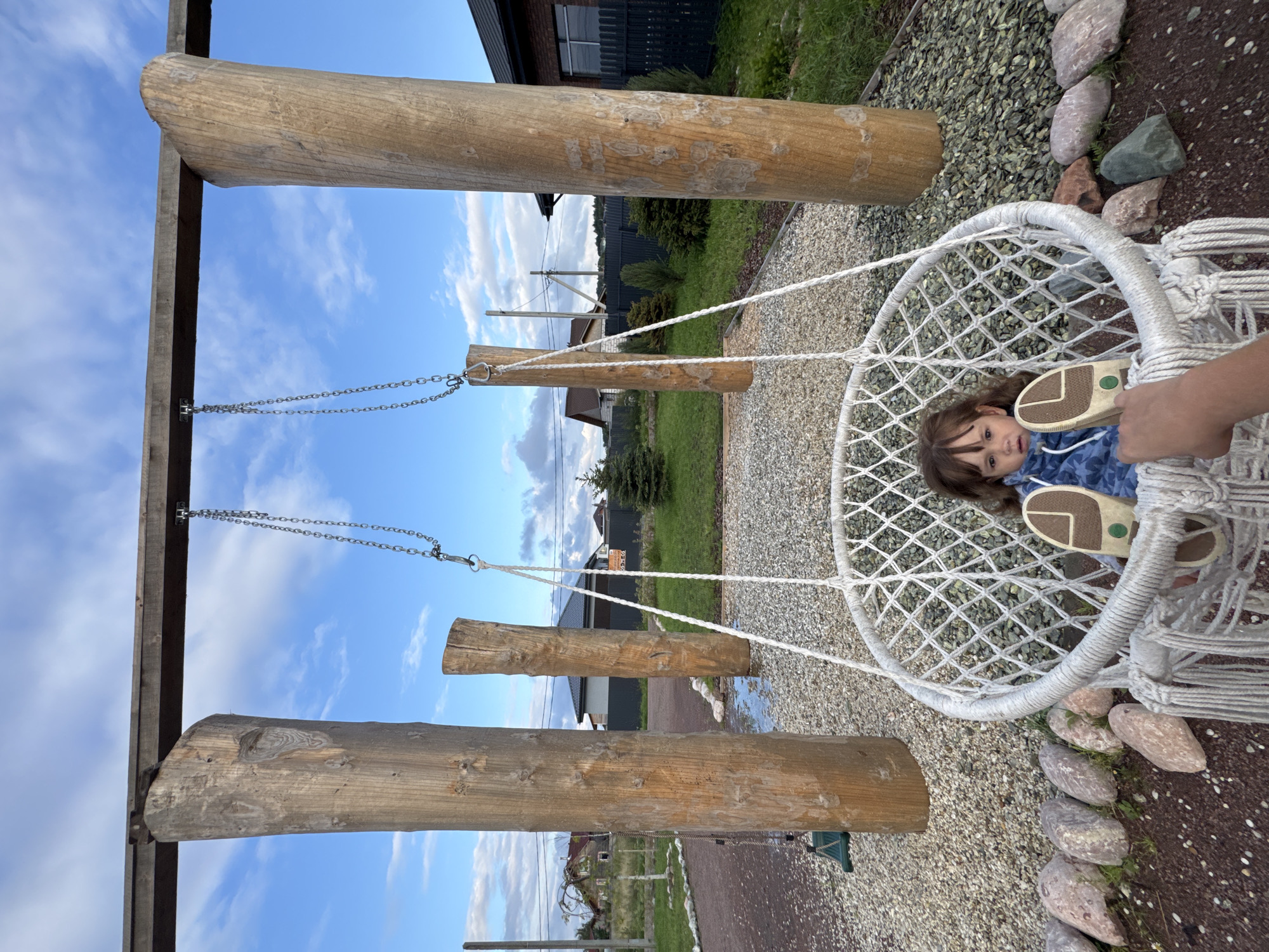 In Timmolovo, Russia, a young girl with brown hair sits in a white macrame hanging chair swing. She is wearing a blue hooded jacket with a star pattern and light-colored shoes. The swing is suspended from a wooden beam supported by two large, rough-hewn wooden posts. The ground around the swing is covered with gravel and a few scattered rocks. In the background, there are houses, a dark fence, and evergreen trees under a cloudy blue sky.