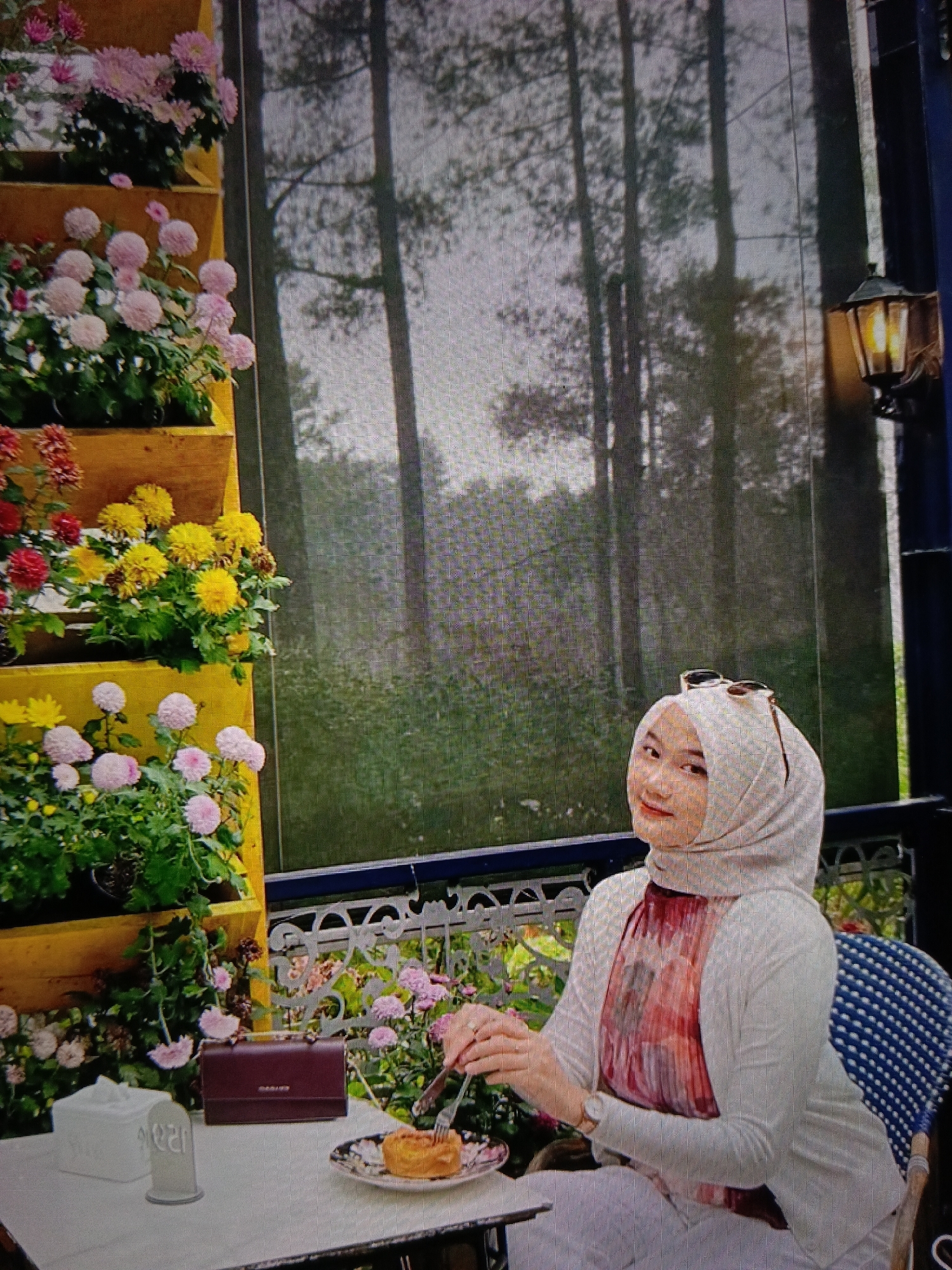 A woman wearing a light-colored hijab and a white cardigan sits at a table outdoors, in front of a vertical planter filled with pink and yellow chrysanthemums. She is smiling and looking at the camera, with sunglasses resting on her head. On the table in front of her, a plate with a pastry and a fork and knife are visible, along with a maroon clutch purse and a white tissue box. Behind the woman, tall trees are visible through a screened enclosure, and a rustic lantern hangs to the right. The scene appears to be in a garden or cafe setting.