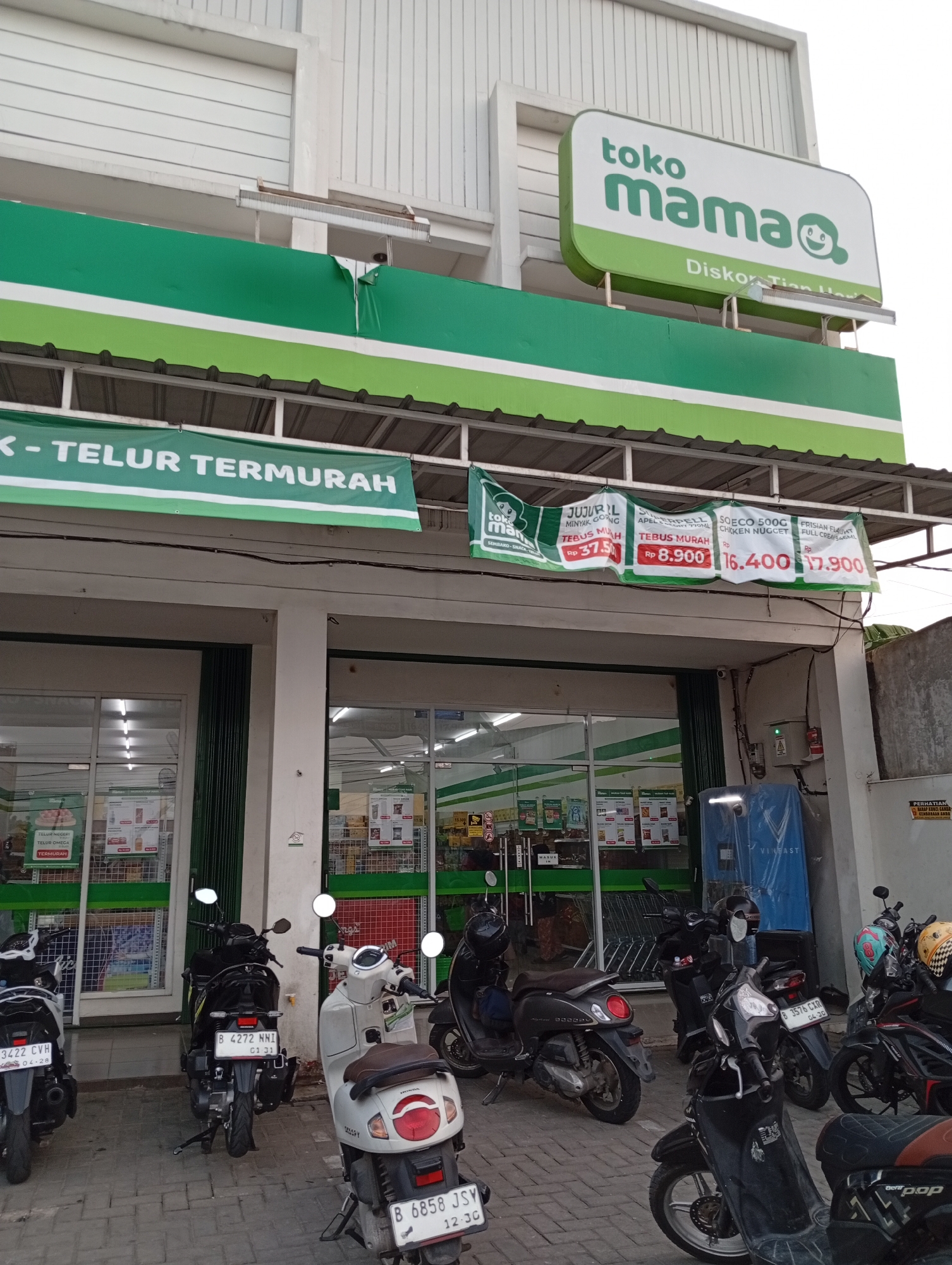 The image shows the exterior of a "Toko Mama" store in Babakan, Indonesia. A large green and white sign above the entrance reads "toko mama" with a cartoon face. Below this, a banner announces "K - TELUR TERMURAH," indicating it's the cheapest egg store. Several promotional banners hang further down, displaying prices for various food items like cooking oil, chicken nuggets, and cookies.
In front of the store, a row of parked motorcycles and scooters are visible, some with helmets on. The store entrance has a glass door and a green metal gate, with trolleys visible inside. The building appears to be a single-story commercial establishment with a white facade and white horizontal siding on the upper portion. The sidewalk is paved with interlocking bricks. A blue charging station for electric vehicles is located to the right of the store entrance. Several security cameras are mounted on the building. The overall scene suggests a busy local market or convenience store.