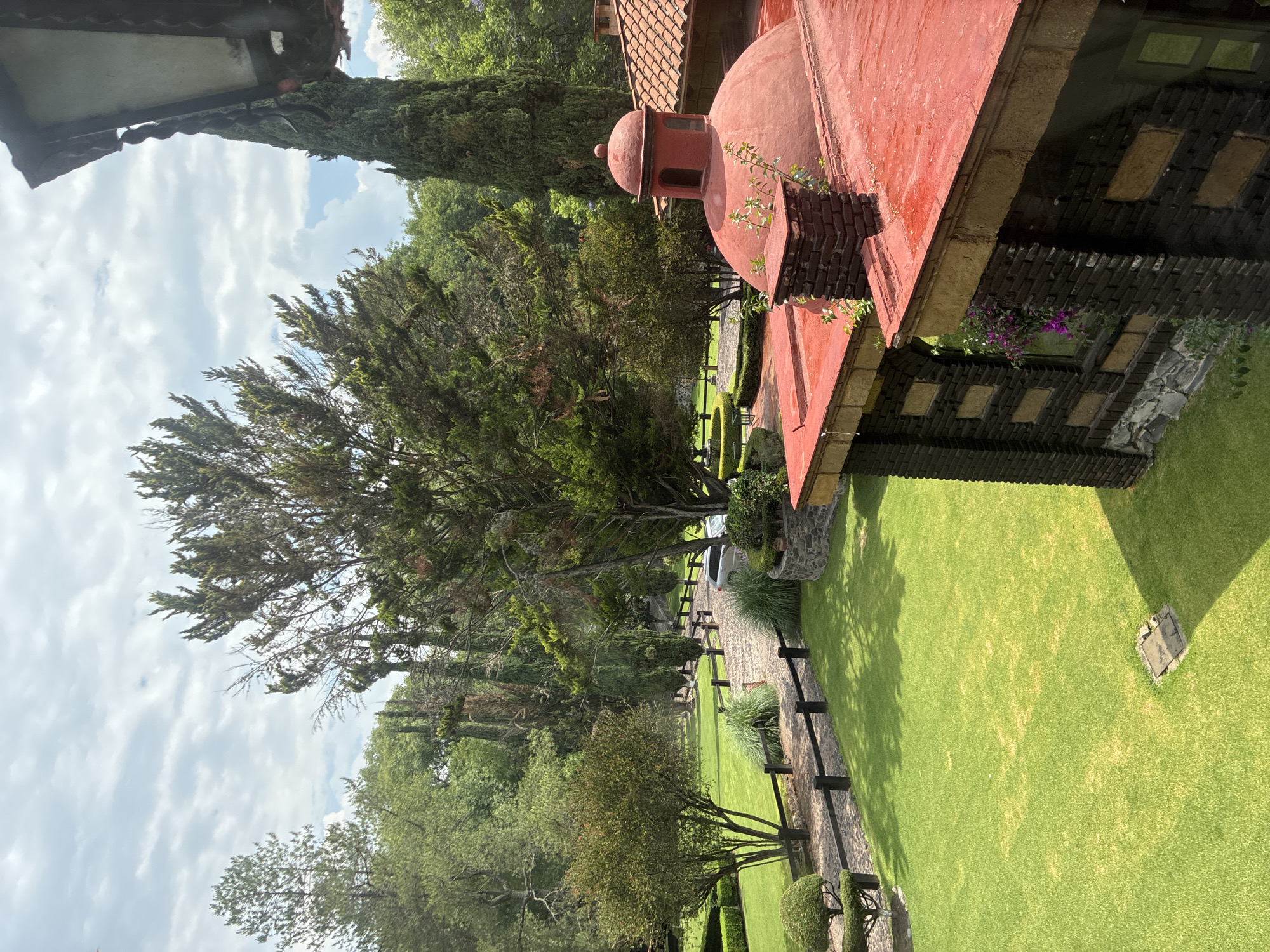 The image depicts a scene in Cañada de Madero, Mexico, featuring a building with a red dome roof, a meticulously manicured lawn, and a tree-lined path. The building's architecture includes dark brown brickwork and stone accents. The lawn is vibrant green and well-maintained. A wooden fence runs alongside a stone pathway lined with trees and hedges. The sky is partly cloudy.