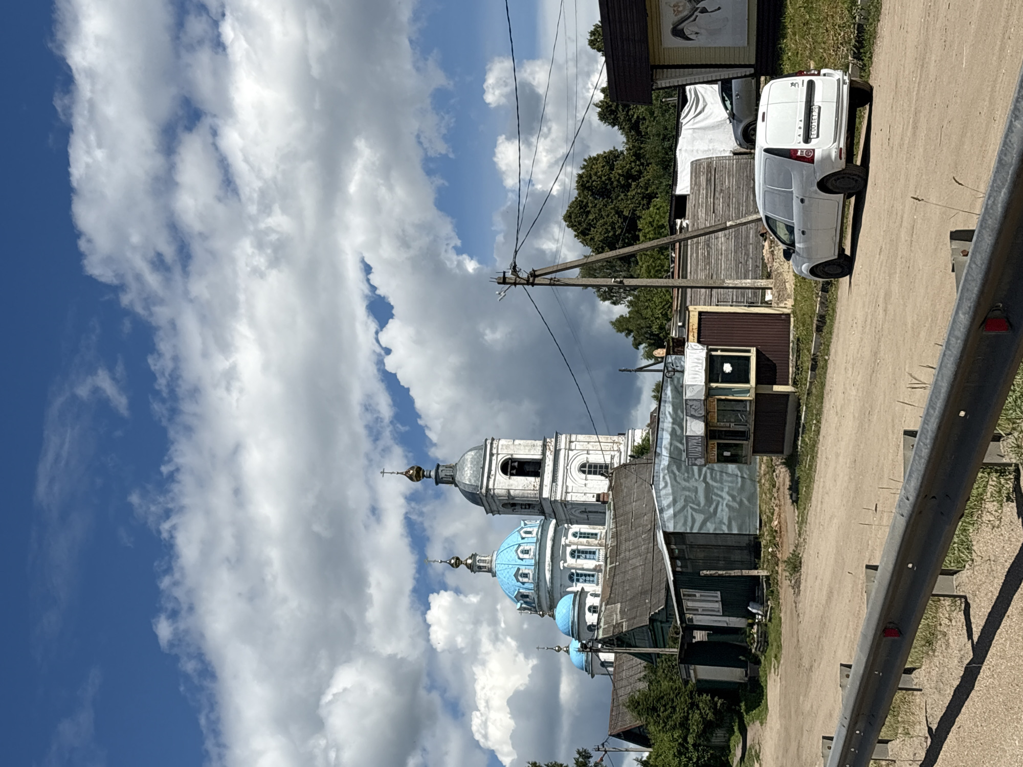 This is a vertical, slightly low-angle photograph of a street scene in a village, likely in Russia, given the provided location context. The primary subjects are a white Lada van parked on a dirt road and a traditional Russian Orthodox church with blue domes. The van is facing away from the camera, and the license plate is partially visible, showing "EOOME 99".
The scene is set on a sunny day with scattered, dramatic white clouds against a bright blue sky. The church, a significant element, features a tall white bell tower with arched windows and a grey dome, topped with a cross. Adjacent to it are smaller buildings, one with distinctive blue domes also topped with crosses. The architecture suggests a historical or religious site.
The road is unpaved and dusty, with a metal guardrail running along the right side of the frame. Several wooden buildings, some appearing weathered and rustic, line the street. The presence of telephone poles and wires indicates infrastructure in the area.
The overall impression is one of a quiet, rural setting on a clear day. There are no people visible in the photograph, and the focus is on the architecture and the natural elements. The lighting suggests it is daytime, likely late morning or afternoon. The image conveys a sense of peacefulness and perhaps a touch of historical charm due to the presence of the church.
