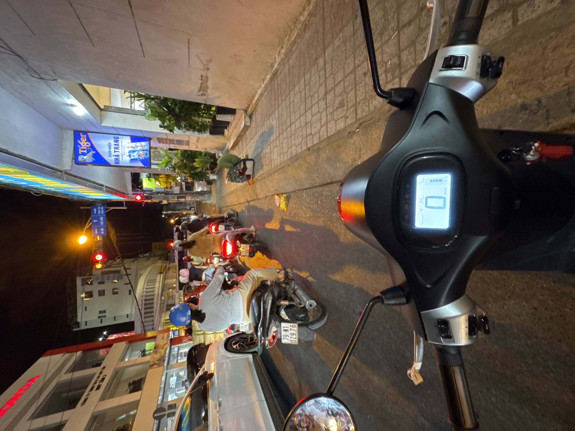 The image captures a nighttime street scene in Nha Trang Ward, Vietnam, as viewed from the perspective of someone riding a scooter. The foreground is dominated by the handlebars and instrument panel of the scooter, displaying a digital readout. In the mid-ground, a traffic jam of motorcycles and cars is visible. Several individuals are on motorcycles, some wearing helmets. A white car is present on the left. The background shows buildings lining the street, one with a "Tiger" beer advertisement. The street is illuminated by artificial lighting, including traffic signals, streetlights, and building lights, casting reflections on the wet pavement. A person is sitting on the sidewalk on the right side of the image. The overall environment suggests an urban setting at night with moderate traffic.