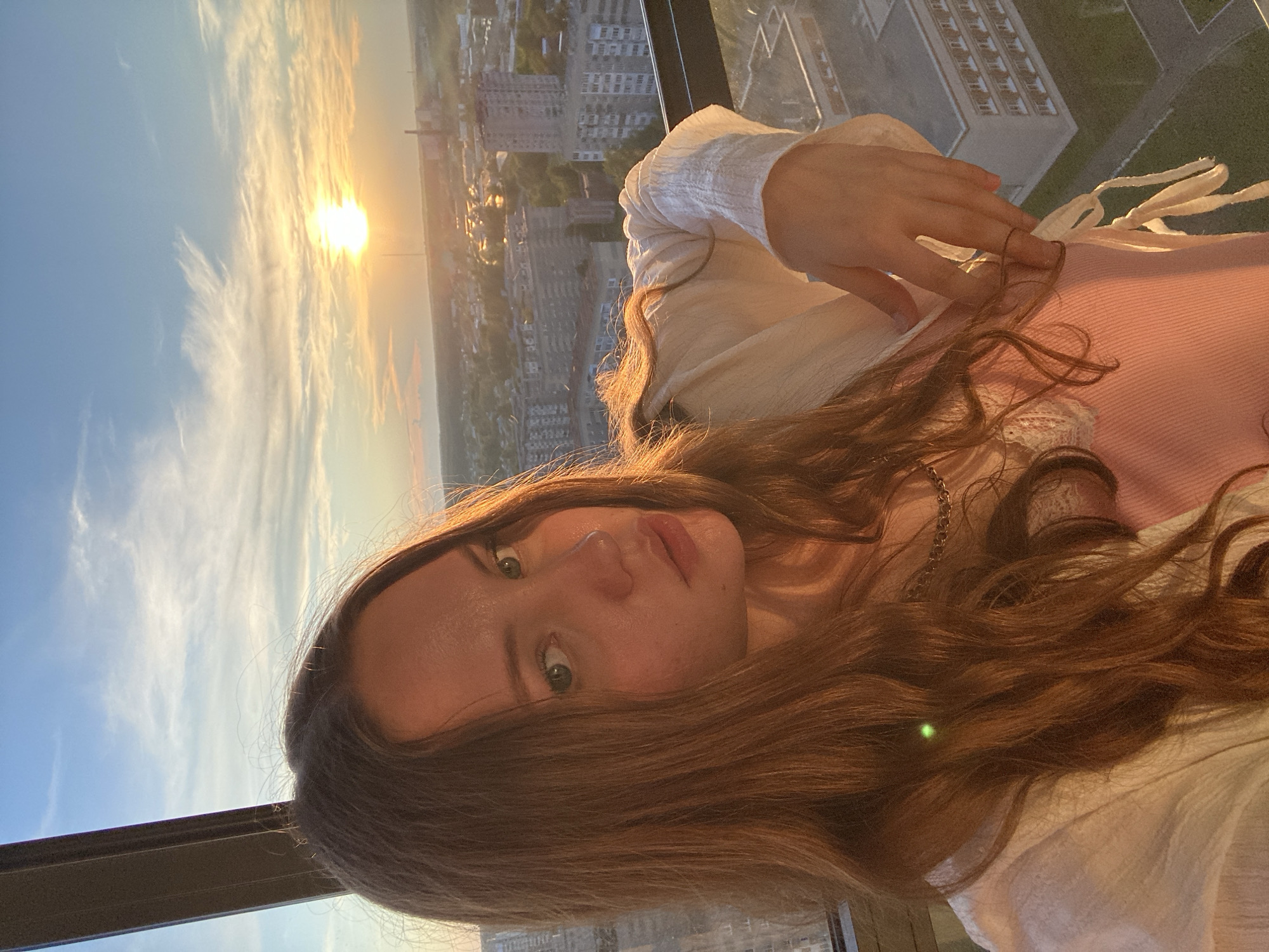 A young woman with long, brown hair is taking a selfie on a balcony overlooking a city at sunset. The woman is wearing a pink tank top and a white, open-front shirt. Her left hand is resting on the white shirt, and her right hand is visible near the edge of the frame. The sun is setting in the background, casting a warm glow over the city. The city appears to be Izhevsk, Russia, with a dense cluster of apartment buildings visible. The sky is blue with streaks of white clouds. The image is framed by a dark structure, possibly part of the balcony or window frame.