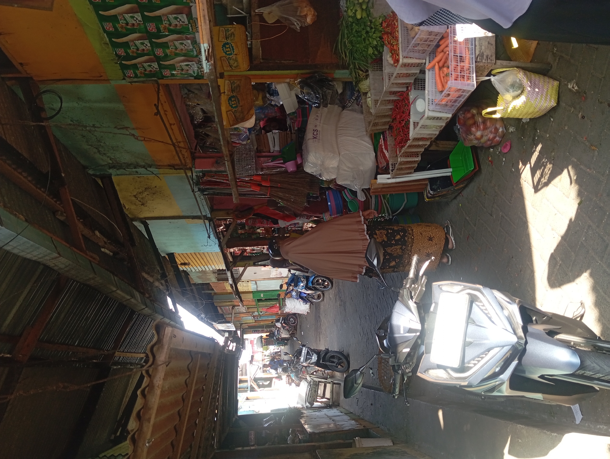 The image depicts a narrow, sunlit alleyway in a marketplace in Kepulungan, Indonesia. The alley is lined with small, colorful stalls selling various goods, including vegetables like chili peppers and carrots, displayed in plastic crates. Brooms and other household items are also visible hanging from the stalls. Several motorcycles are parked along the alley, and a woman wearing a headscarf and a patterned skirt is walking away from the camera. The ground is paved with cobblestones, and the overall atmosphere suggests a bustling local market on a bright day. The structures appear to be made of corrugated metal and wood, with some showing signs of age and weathering.
