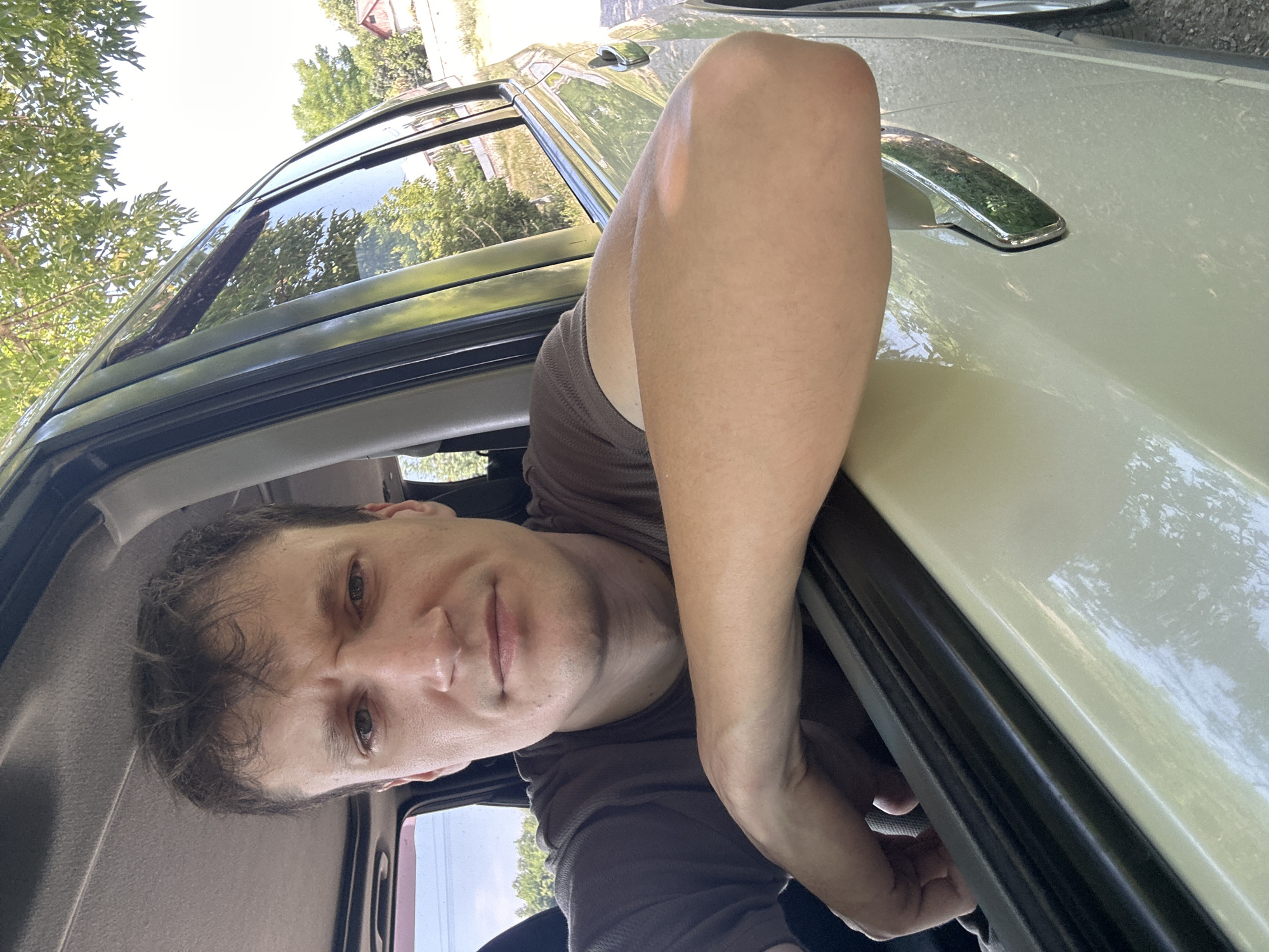 A man with short, brown hair is leaning out of the open window of a light-colored vehicle, resting his forearm on the car door. He is wearing a brown t-shirt and appears to be looking directly at the camera. The car is outdoors, with a tree and foliage visible in the background, suggesting a rural or suburban setting. The overall lighting indicates it is daytime.