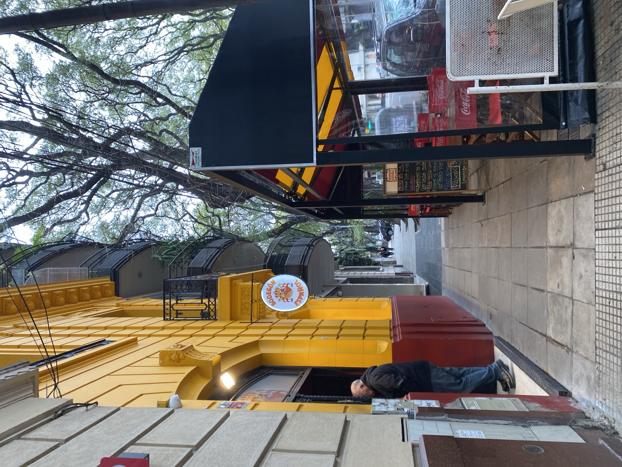 The image shows a sidewalk scene in Buenos Aires, Argentina. A man in a dark jacket and jeans stands near the entrance of a yellow building with red trim. A circular sign hanging from the building reads "BODEGON ESPAÑOL". To the right of the building, an outdoor seating area for a restaurant is set up with tables and chairs under a black awning with yellow and red accents. The tables are covered with clear plastic. A menu is visible on a blackboard in the seating area. Trees with lush foliage line the street, and their branches extend over the sidewalk. Cars are parked along the street. The architecture suggests a residential or commercial area in a city.