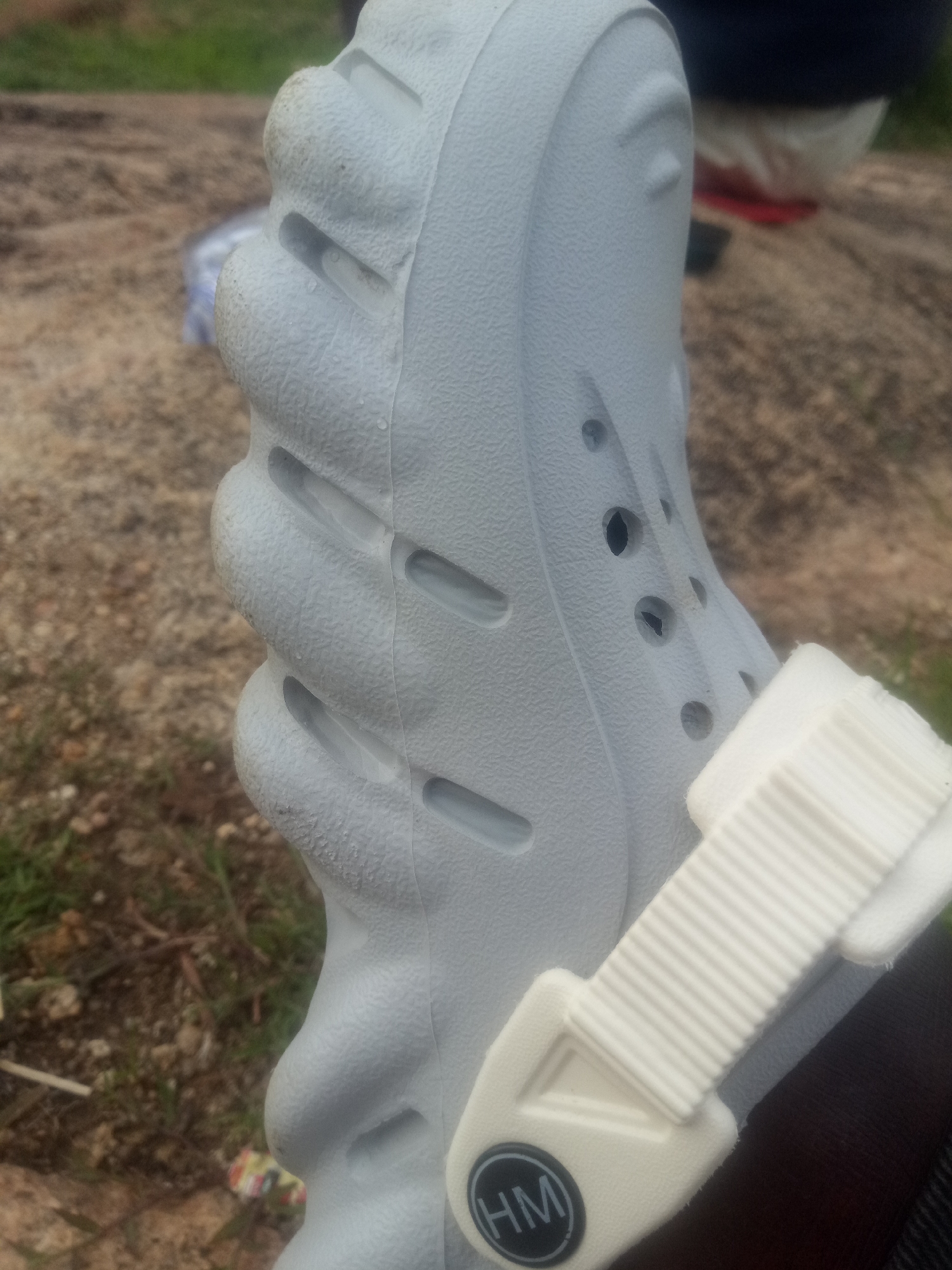 The image shows a close-up of a light gray shoe with a white strap. The shoe has a textured surface and several circular holes. The strap is ribbed and attached to the shoe with a triangular piece. A black circular emblem with the letters "HM" in white is visible on the strap. The shoe appears to be made of a lightweight material like rubber or foam.
The background is blurred and appears to be an outdoor setting with rough, rocky ground and some green vegetation. There are also some out-of-focus objects in the background, including what looks like a plastic bottle and some colorful debris. The lighting suggests it might be daytime, possibly overcast.
The image is focused on the shoe, suggesting that the shoe is the primary subject. There are no people clearly visible, but a dark-colored leg or foot is partially visible beneath the strap, indicating that the shoe is being worn. The activity taking place is simply the shoe being presented or observed in an outdoor environment. The overall impression is one of casual footwear in a natural or uncultivated setting.