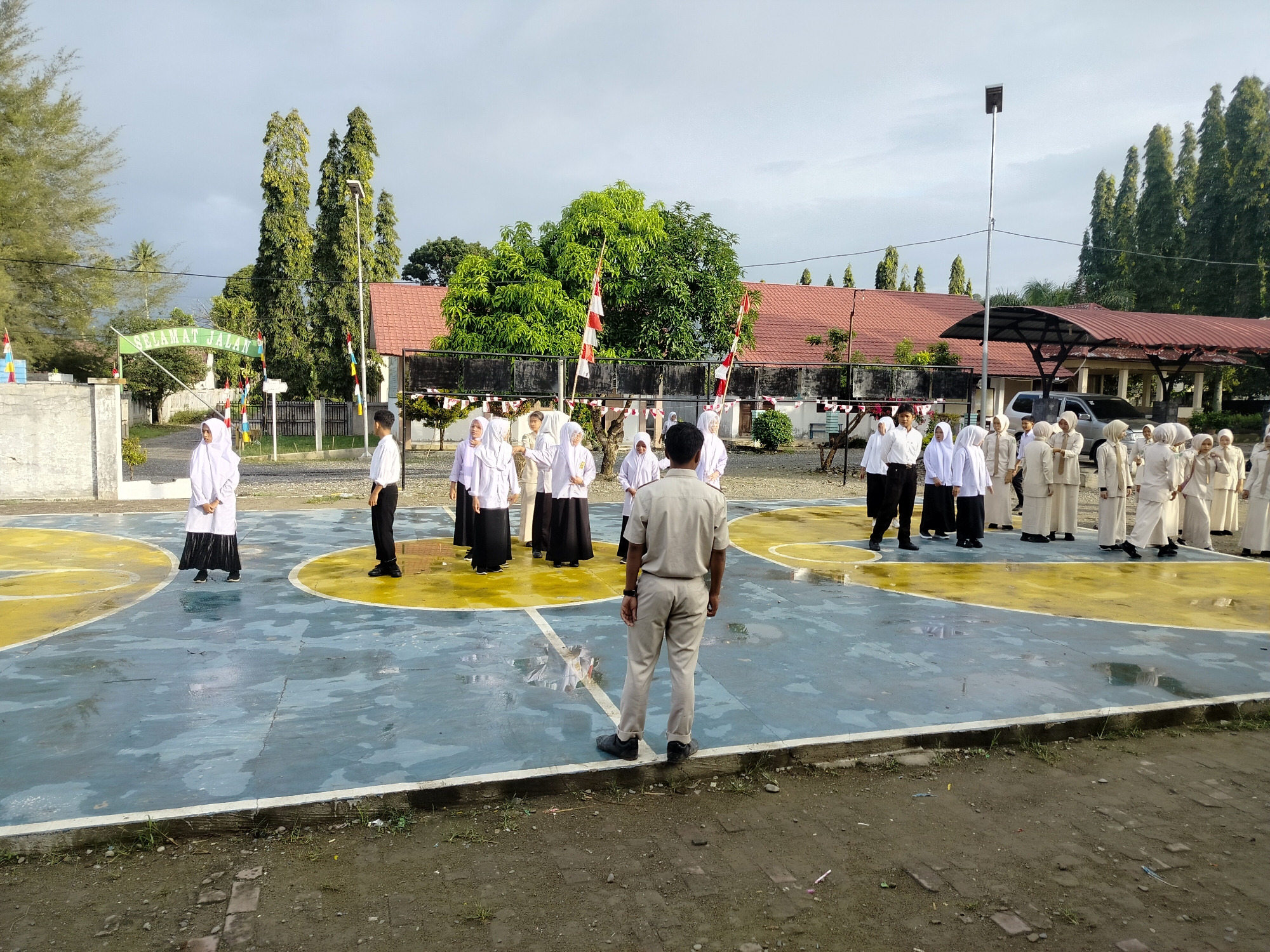 A group of students wearing white uniforms and headscarves are assembled on a wet basketball court. The court has blue and yellow markings, and there are puddles of water on its surface. In the background, a building with a red tiled roof is visible, along with several tall, dark green trees. Flags are displayed on poles in front of the building. A man in a light brown uniform and pants is standing on the court, facing away from the camera. Another person in a white shirt and black pants is standing to the left of the man in uniform. The sky is overcast, suggesting it may have recently rained or will rain soon. The scene appears to be outdoors at a school or educational facility.