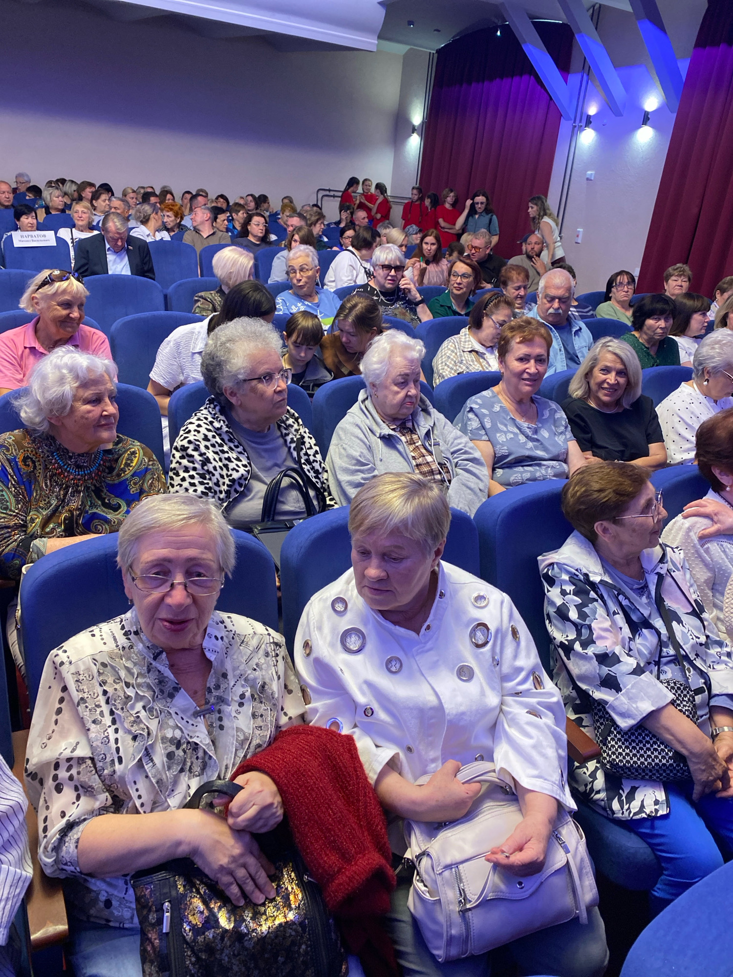 A large group of people, predominantly elderly women, are seated in blue upholstered chairs in an indoor venue, likely a theater or auditorium. The room is dimly lit, with overhead lights casting a blue hue on the walls and ceiling. The audience members are facing forward, presumably towards a stage or screen that is not visible in the frame. Many of the women are dressed in casual attire, with some wearing patterned blouses and others in solid colored tops. Several individuals are holding handbags, and one woman in the foreground has a red knitted shawl draped over her arm. The overall atmosphere appears calm and attentive. The scene is located in the city of Ivanteyevka, Russia.