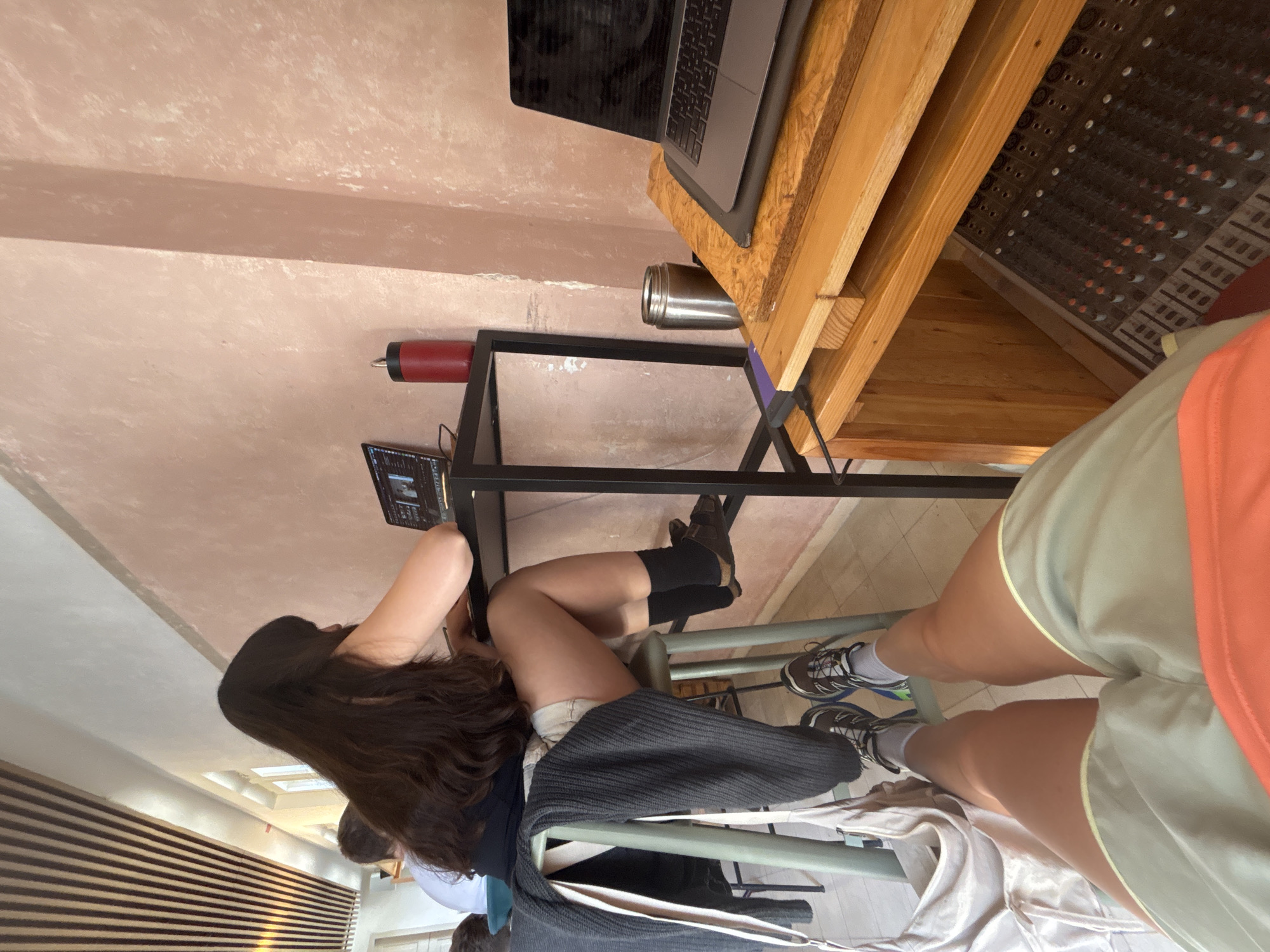 An indoor scene, possibly an office, studio, or collaborative workspace, captured from a slightly elevated and tilted perspective. The image features two individuals.
In the foreground and to the left, a person with long, dark hair is seated at a table, intently looking at and interacting with a small laptop or tablet. They are wearing light-colored shorts and dark socks with boots, and a dark sweater or jacket is draped over a chair. Their arms are visible as they operate the device.
To the right, another person is partially visible, primarily showing their legs and lower torso. They are wearing light green shorts with a yellow stripe, an orange top, and athletic shoes with white socks. This person appears to be standing or sitting close to the working area.
The environment includes a black metal frame table or stand where the first person's laptop rests. Above this, a larger laptop is open on a wooden stand, alongside a silver tumbler and a red bottle. To the far right, a wooden structure houses what appears to be a sound mixing board or control panel with numerous buttons and faders. The walls are a light, textured, possibly pinkish-beige color, with some white mottling and a prominent horizontal beam. Part of a wall with horizontal wooden slats is visible in the background, along with what might be skylights or ceiling lights.
The lighting suggests it is daytime. No specific emotions are discernible, and no legible text is visible in the image. The location context provided is Tel-Aviv, Israel.