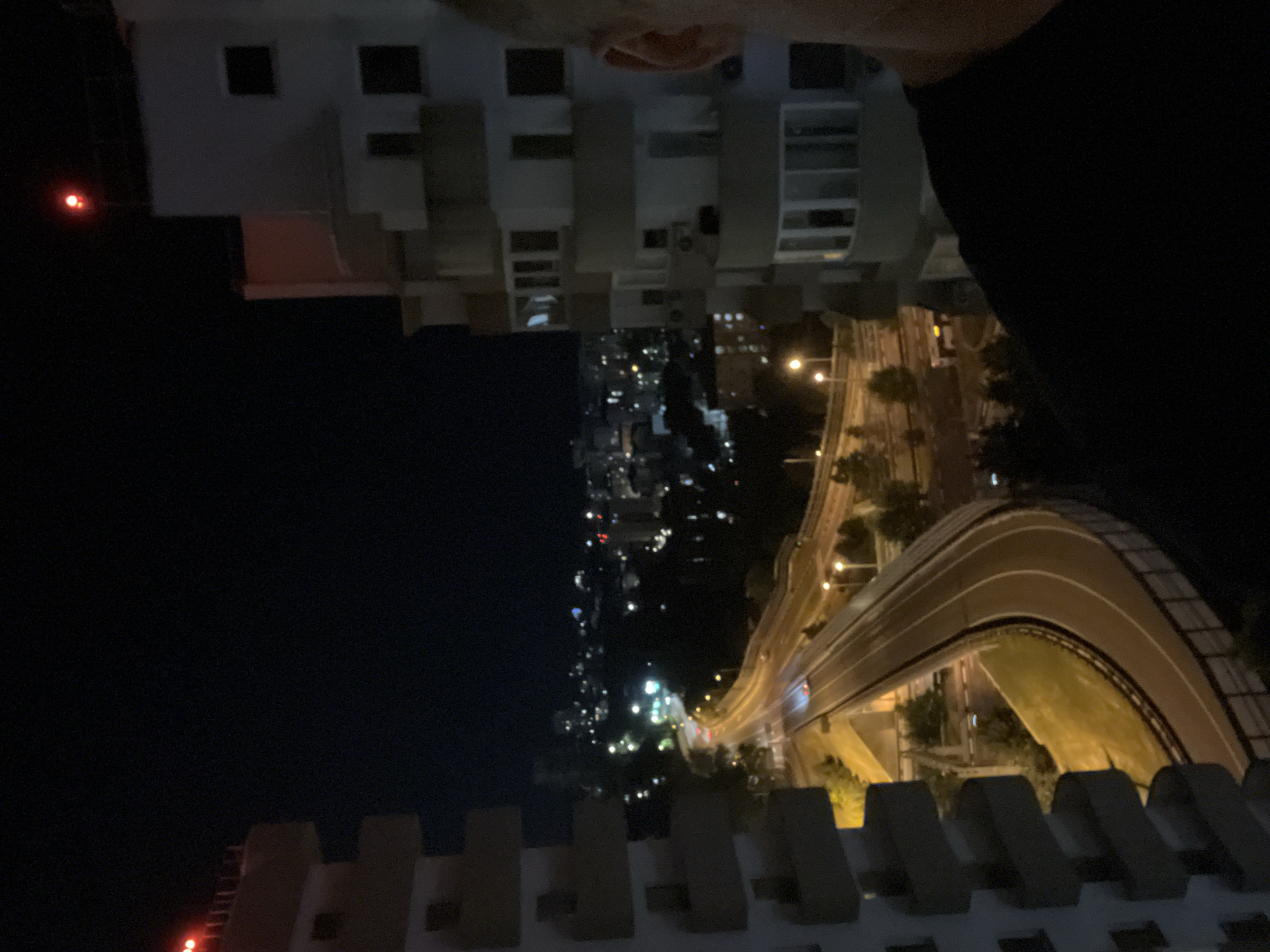 An outdoor, nighttime, long-shot, eye-level, medium-wide, horizontal, full-color, slightly low-contrast photograph shows an urban scene in Sochi, Russia, with a street and buildings illuminated by artificial lights. In the foreground, on the right, the shoulder and side of a person wearing a black shirt are visible. On the left, the side of a large, multi-story building with numerous balconies is visible. A curved roadway with streetlights and a few cars is in the mid-ground, winding through a landscape dotted with trees and other buildings, some of which are illuminated. In the far distance, more buildings form a cityscape under a dark, clear sky. Two red lights are visible at the top of two buildings, one on the left and one on the right.