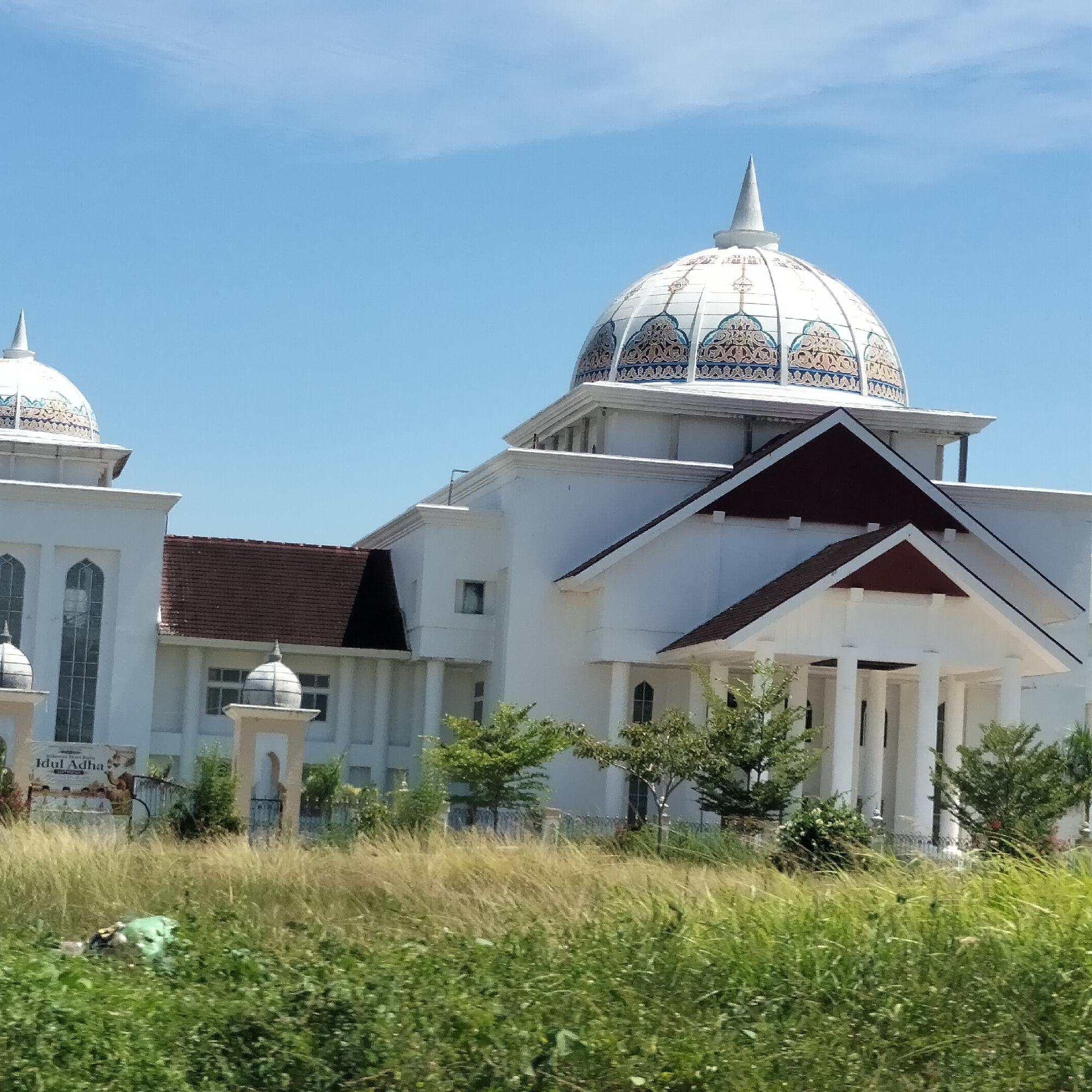 The image captures a white building with a decorative dome in Meunasahaluedrienlhoksukon Barat, Indonesia. The dome is white with ornate patterns and a pointed spire. The building has a tiled roof and a portico with white columns. In front of the building is overgrown grassy vegetation, partially obscuring a sign that reads "Idul Adha". The sky is clear blue.