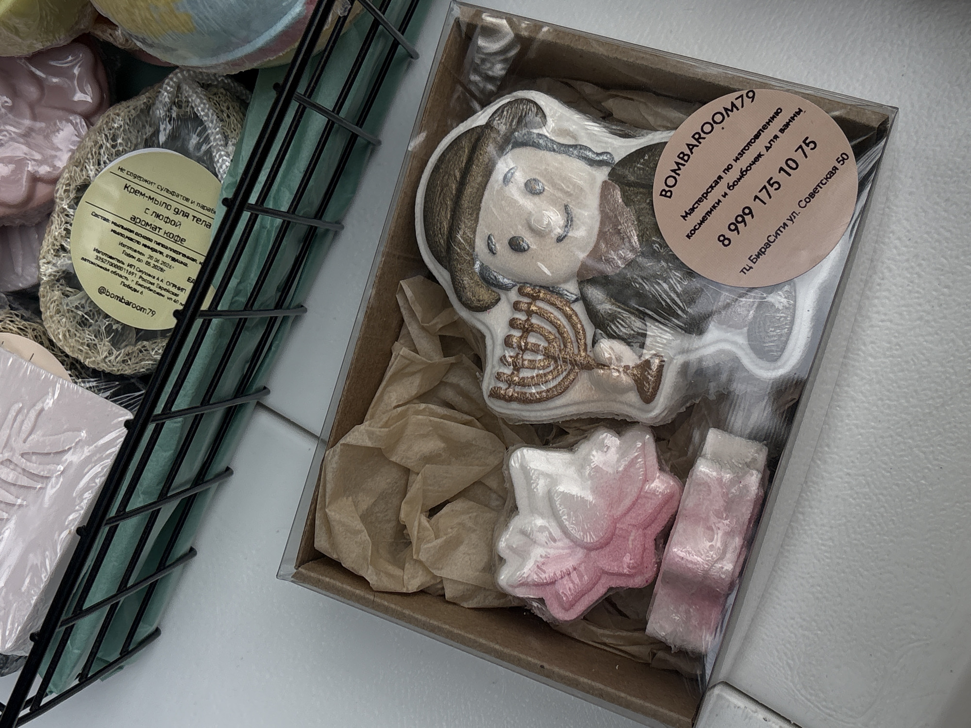 An overhead shot shows a clear plastic-wrapped gift box containing bath bombs. The box, resting on a white surface, is filled with crumpled brown paper. Inside, there are three bath bombs: a pink and white star-shaped bomb, a small pink rectangular bomb, and a large white bath bomb shaped like a person in a hat holding a menorah. The bath bomb person has a dark hat, brown hair, and is wearing a dark green robe. A round, light pink sticker is affixed to the plastic wrap of the gift box. The sticker features text in Russian. The text includes "BOMBAROOM79" at the top, followed by "Мастерская по изготовлению косметики и бомбочек для ванны" (Workshop for the production of cosmetics and bath bombs). Below that is the phone number "8 999 175 10 75". At the bottom of the sticker is "ТЦ БираСити ул. Советская 50" (TC Biracity, Sovetskaya St. 50), indicating the location in Birobidzhan, Russia. In the upper left of the image, a portion of a black wire basket containing more bath products is visible.