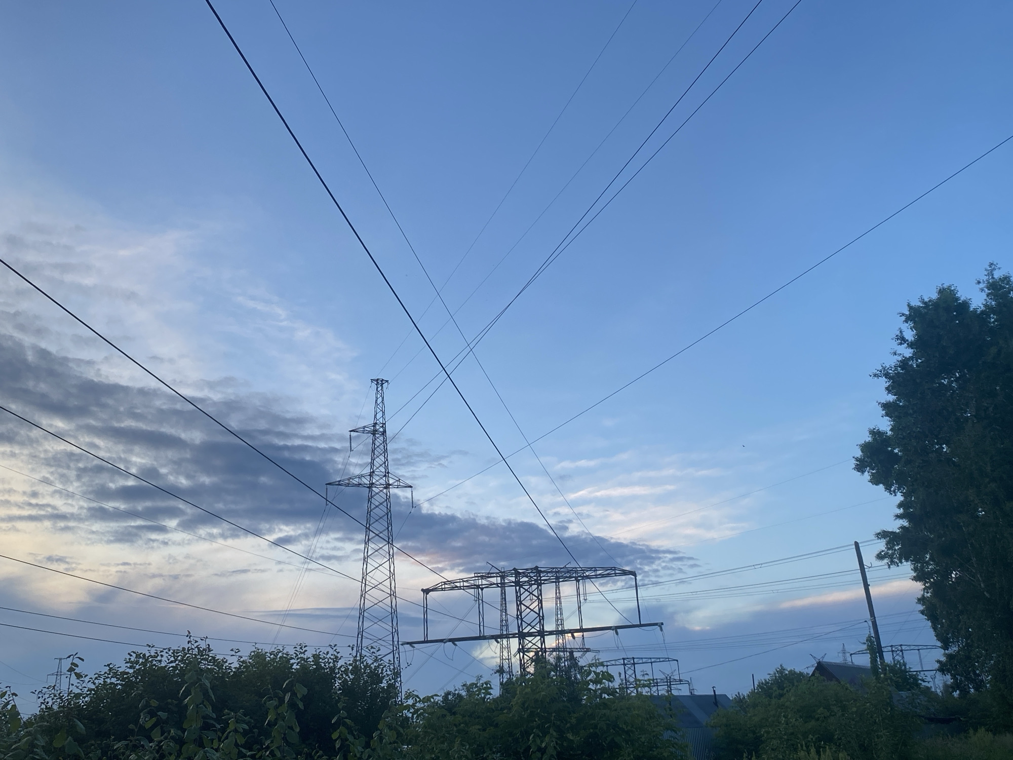 In an outdoor scene, power lines crisscross against a twilight sky filled with a mix of clouds and clear blue. The sky exhibits a gradient of blues, with hints of pink and yellow near the horizon where the sun has set. In the foreground, lush green foliage forms a dense canopy. Towering electrical pylons, constructed from dark metal lattice, dominate the midground and background, their skeletal structures silhouetted against the sky. One prominent pylon stands tall on the left, while others of varying heights and designs are visible further back. The power lines themselves, thin black strands, are strung between these structures and extend across the frame, creating a complex network against the open sky. The overall impression is one of infrastructure set against a natural backdrop, captured during the transitional light of dusk.