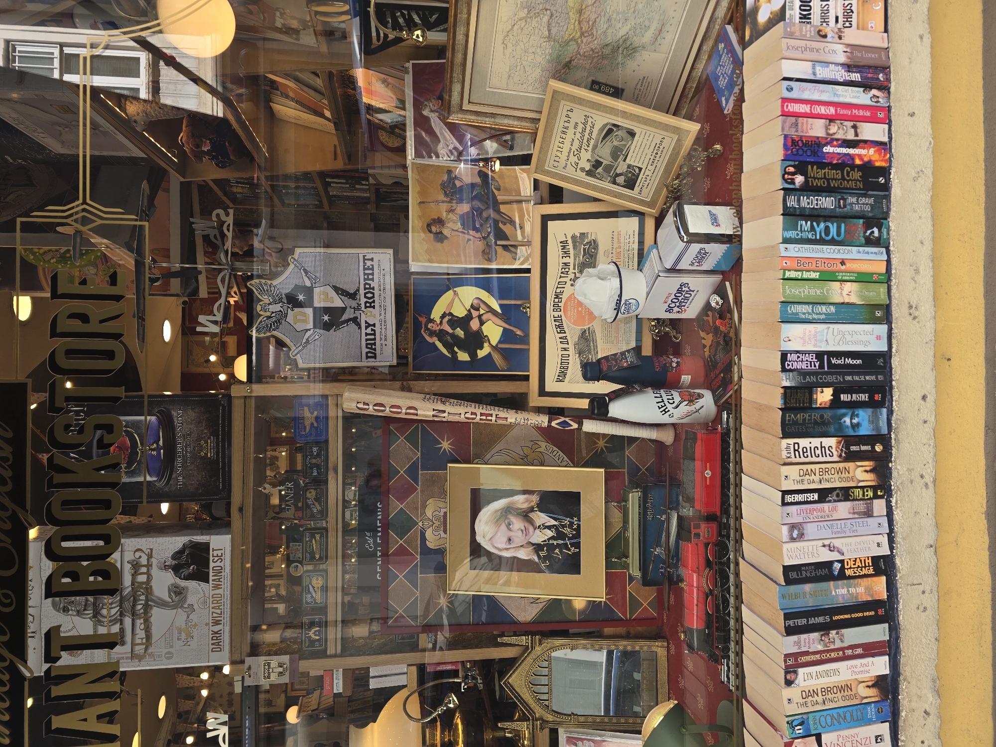 This image captures a forensic view of the display window of the Elephant Bookstore in Sofia, Bulgaria. The glass frontage prominently features large, gold-colored cutout letters spelling "ELEPHANT BOOKSTORE" across the top. Reflections on the glass include parts of an exterior building with a white window and vertical blinds, along with warm interior and exterior light sources.
The display itself is multi-layered. In the upper section, various merchandise boxes are visible, including a "DARK WIZARD WAND SET" box depicting Voldemort, and a "THE SORCERER'S STONE PROP REPLICA" box. A "DAILY PROPHET" banner with an owl emblem is also affixed to the glass.
The central display area features a gold-framed, autographed photograph of actress Evanna Lynch (Luna Lovegood), positioned against a backdrop of a shield-like crest design in blue, red, and gold. To the left of the photograph, a small, ornate gold-colored mirror and a brass desk lamp are visible. To the right, a wooden baseball bat inscribed with "GOOD NIGHT" and other text vertically, alongside a white and a dark "HELLFIRE CLUB" branded bottle, are arranged. Behind these items, several framed vintage-style posters are displayed, depicting a witch on a broomstick and a woman on a carousel horse. Further right, a framed antique map and two framed advertisements, one for a "Studebaker" car and another in Cyrillic script, are present.
The frontmost part of the display comprises a horizontal row of numerous paperback books, spines facing forward, resting on a ledge. Notable titles include "The Da Vinci Code," "Death Message," and "Wild Justice." Behind this row of books, a red model train, resembling a steam locomotive with its tender, is positioned on tracks. To the right of the train, "SCOOPS AHOY" branded items, including a white cup, a cup holder, and a dispenser, are visible. The entire display surface is covered with a red, textured fabric.
