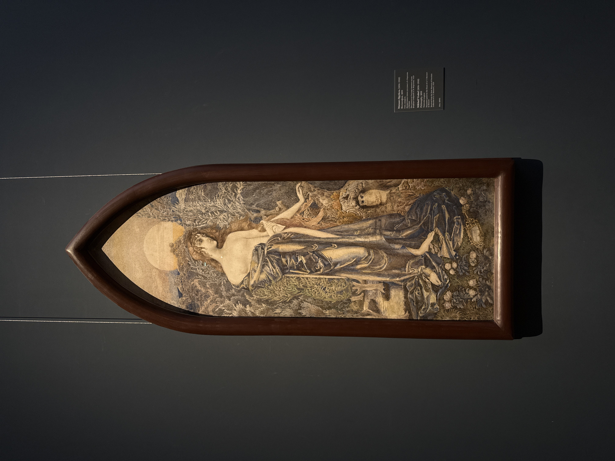 The image features a painting, titled "Philosophy" by Mikhail Vrubel, displayed on a dark grey wall. The painting is encased in a wooden frame with a unique, peaked top. The artwork depicts a nude woman lying horizontally, reaching towards a skeleton. The scene is set against a backdrop of greenery, with a light circle behind the woman's head. A small placard with text regarding the artwork and artist is positioned to the right of the painting on the wall. The image appears to be taken in a museum setting, presumably in Moscow, Russia.