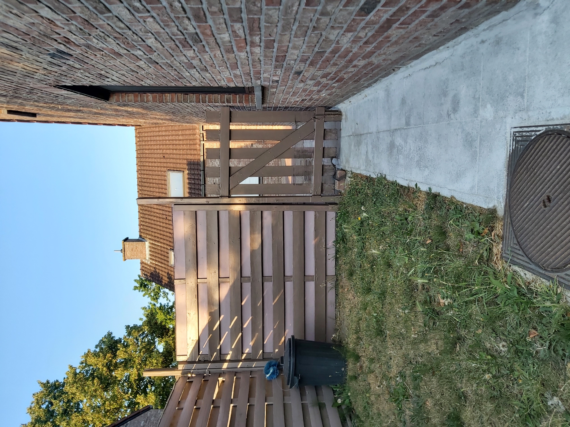 The image depicts a narrow backyard alleyway adjacent to a brick building. On the left, a wooden fence with horizontal slats is visible, extending to a wooden gate. A dark green trash can with a blue cleaning brush rests near the fence. The ground is covered in patchy grass and weeds. A concrete walkway runs alongside the brick building on the right, leading towards the gate. The building features a dark window and a red brick facade. In the background, a tiled roof with a chimney and trees are visible under a clear blue sky. The scene is illuminated by natural sunlight, casting shadows from the fence onto the grass and gate. The overall impression is that of a utilitarian, somewhat unkempt, outdoor space.