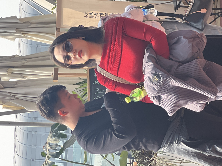 A man and woman stand outdoors. The woman wears a red sweater and sunglasses, a cream-colored purse strap over her shoulder. A grey jacket partially obscures her. The man wears a black shirt. A green bottle sits near them. In the background, there are umbrellas, plants, and a building.