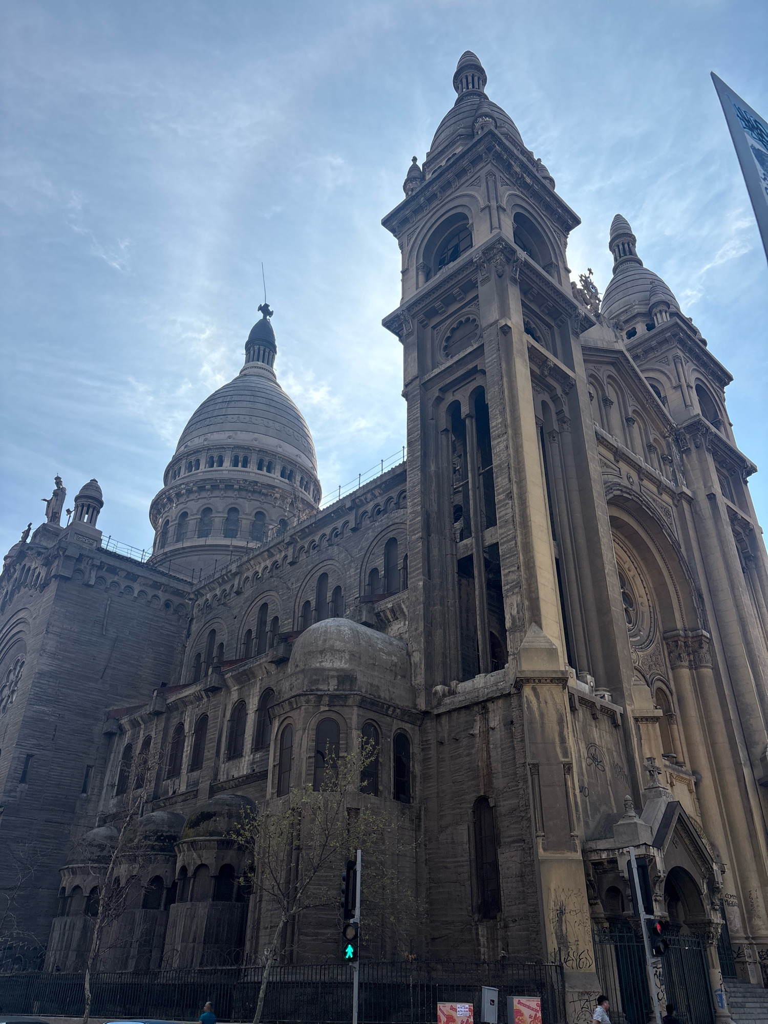 This low-angle, wide shot captures the imposing facade of the Basilica de la Gratitud Nacional in Santiago, Chile, under a partly cloudy sky. The architecture is characterized by its Romanesque Revival style, featuring intricate stonework, arched windows, and multiple domes and spires. The building appears to be constructed from a light-colored stone that has weathered over time, with some areas showing signs of discoloration or staining.
Prominently in the foreground, to the right, is a traffic light displaying a green signal, with a pedestrian crossing signal showing a walking figure. In the mid-ground, a black metal fence runs along the perimeter of the building, and beyond it, a few bare trees are visible. A couple of people are present near the fence, one walking and another appearing to be standing. There are also some posters or advertisements affixed to structures near the fence.
The sky is a mix of blue and white, with streaks of clouds, and the sun is partially obscured by a building, creating a bright halo effect. The overall impression is one of grandeur and historical significance, with a sense of urban life surrounding the religious structure.