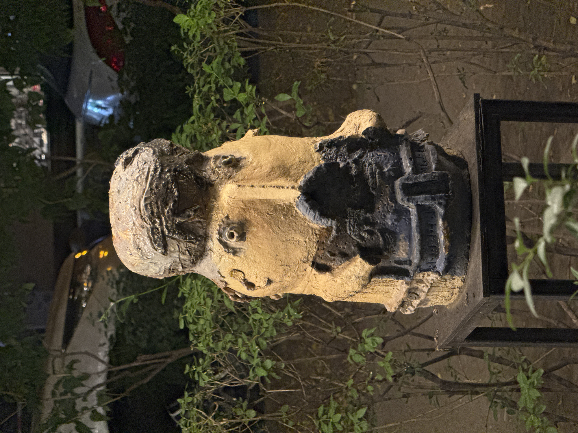 A statue of a head rests on a low platform, likely a plinth. The head is partially painted, the majority being a yellowish color, with blue and black markings on the top of the head and the right side of the face. The statue is located outdoors, surrounded by greenery. In the background, there are indications of streetlights, a road and some buildings, but the surroundings are blurry. The statue is in Yerevan, Armenia.