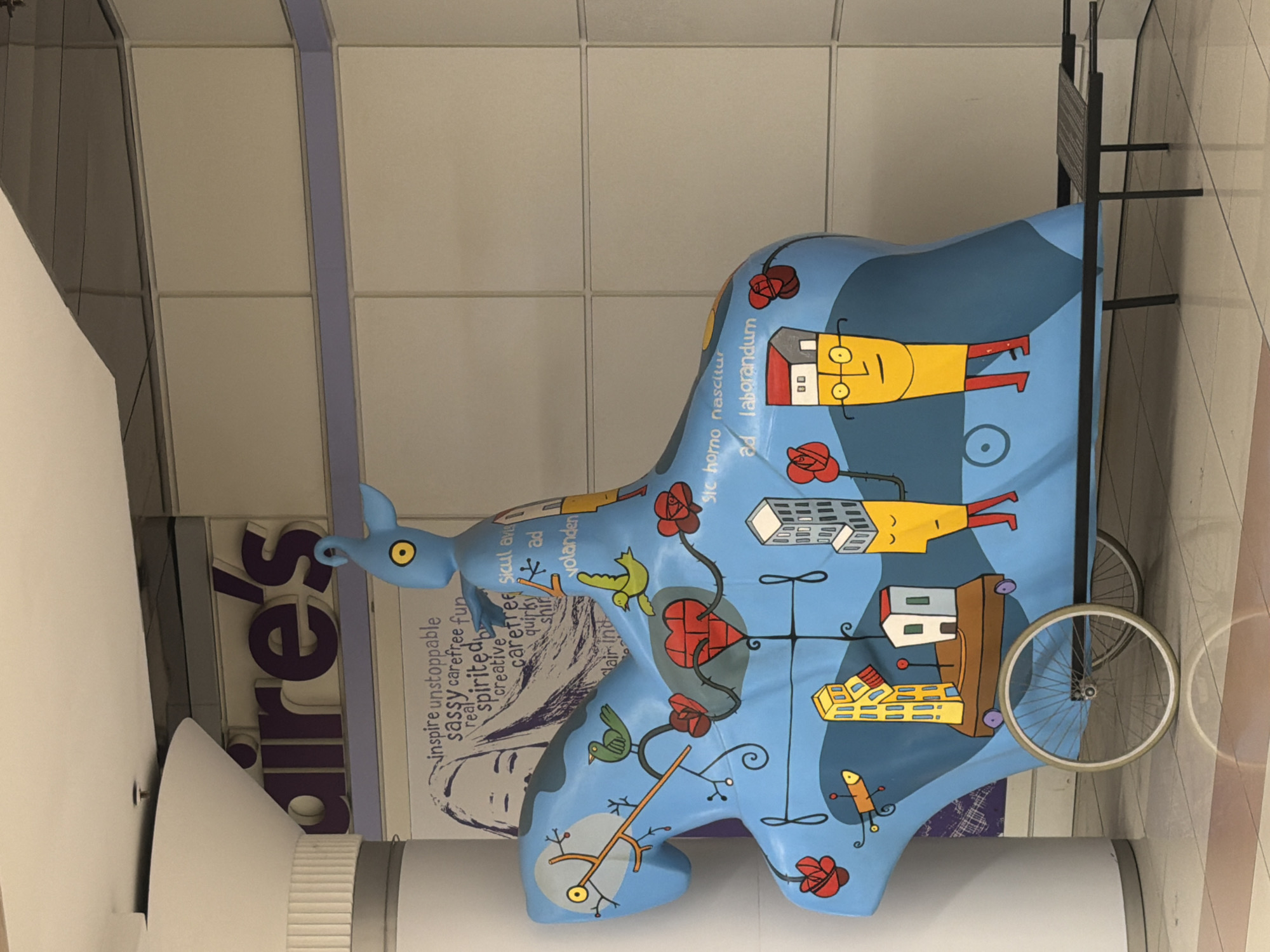 This is a vertically oriented photograph of a brightly painted, blue, elephant-shaped sculpture. The sculpture is adorned with cartoonish drawings, including red roses, birds, hearts, and abstract figures. Some parts of the sculpture resemble buildings with windows, and one figure appears to be a yellow, rectangular head with glasses and red legs, suggesting a human or humanoid representation. The sculpture has wheels, implying it might be a vehicle or meant to evoke transportation.
In the background, there are large, purple letters spelling out "aires." Above and behind the sculpture, a grid of cream-colored tiles forms the wall or ceiling of the setting, with a thin purple band running vertically. A dark metal structure, possibly part of a bench or display, is visible to the right of the sculpture. The lighting suggests an indoor environment, possibly a mall or public space.
Text visible on the sculpture includes phrases like "inspire unstoppable," "sassy carefree fun," "respirited," "creative," "carefree soul," "quirky," and Latin phrases such as "ste horno nascitur" and "ad laborandum." The overall impression is whimsical and artistic, with the colorful decorations and playful figures creating a cheerful and engaging scene. The photograph is taken from a slightly elevated perspective, looking down on the sculpture.