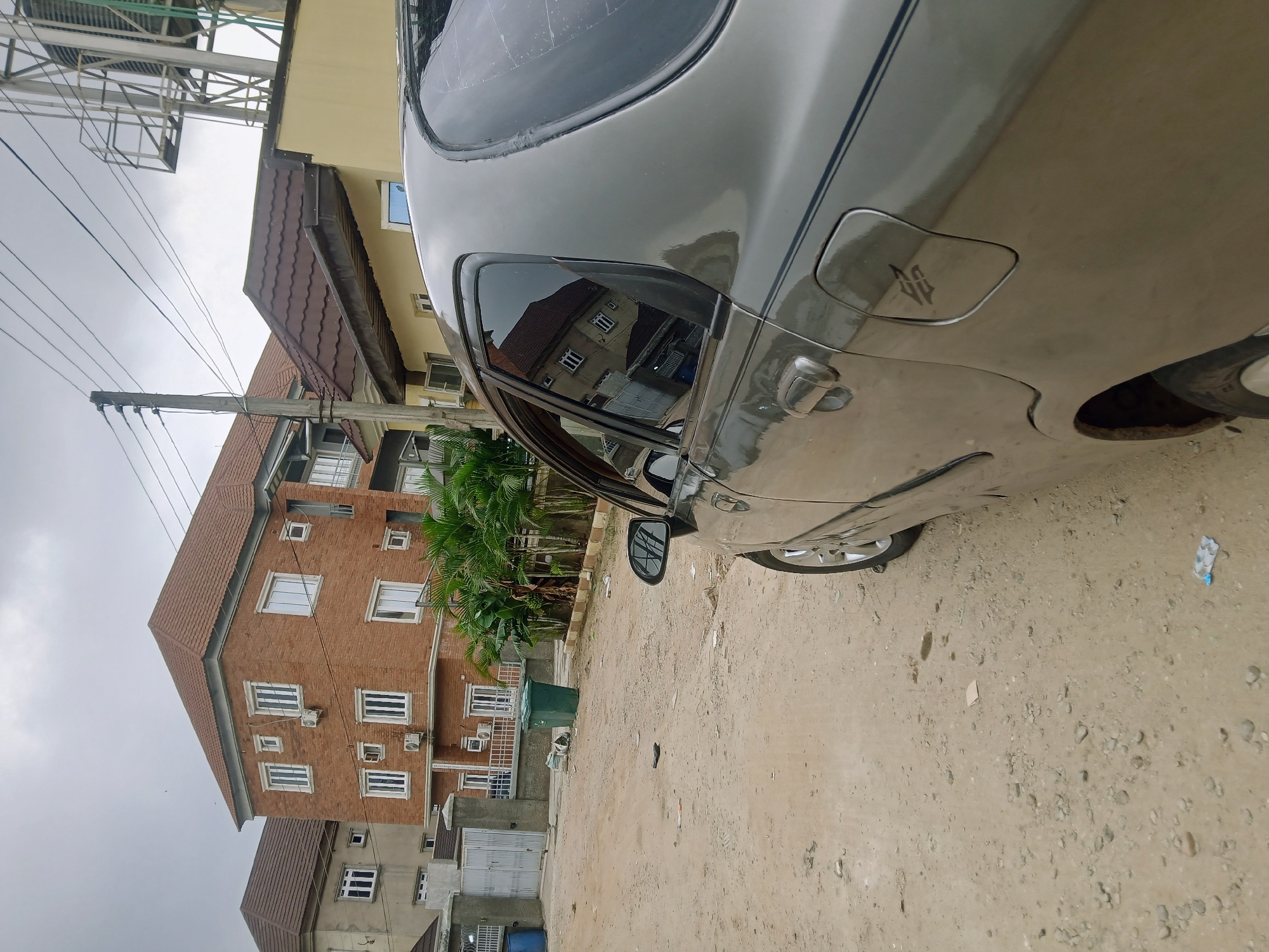 The image shows a portion of a gray car parked on a dirt road in Jiru, Nigeria. Adjacent to the road is a building complex with brick and yellow-painted sections, featuring windows, balconies, and a red-tiled roof. Palm trees and a gate are visible in front of the building. Overhead, there are utility poles and wires. The sky appears overcast.