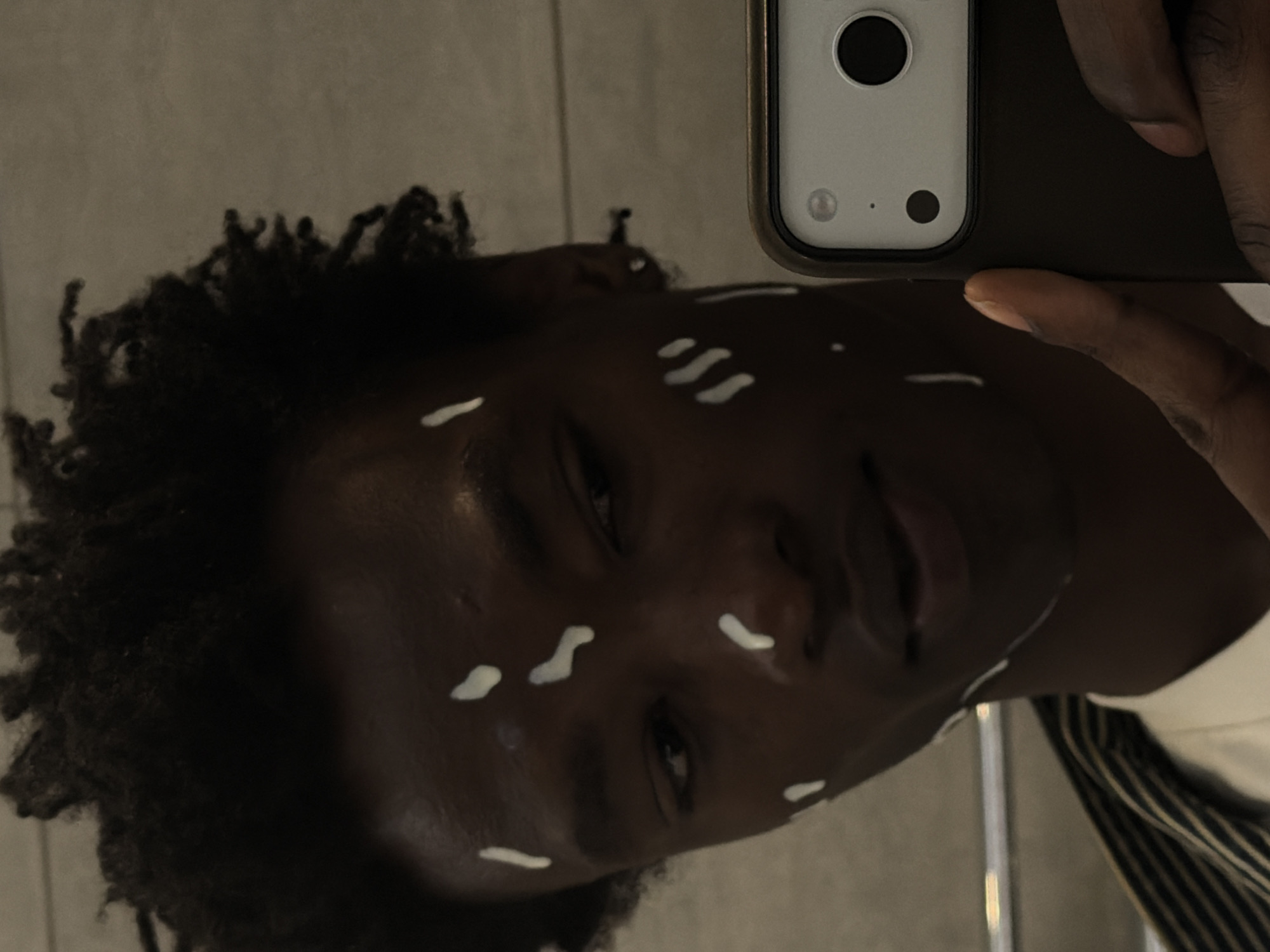 A dark-skinned person with short, dark, coiled hair takes a selfie in front of a light grey wall. The person is holding a silver and black smartphone in their right hand. They have several white, curved lines of cream or lotion on their face. The person's head is tilted slightly to the left. A white shirt with narrow, dark stripes is visible at the bottom of the frame. No evidence of a London location.