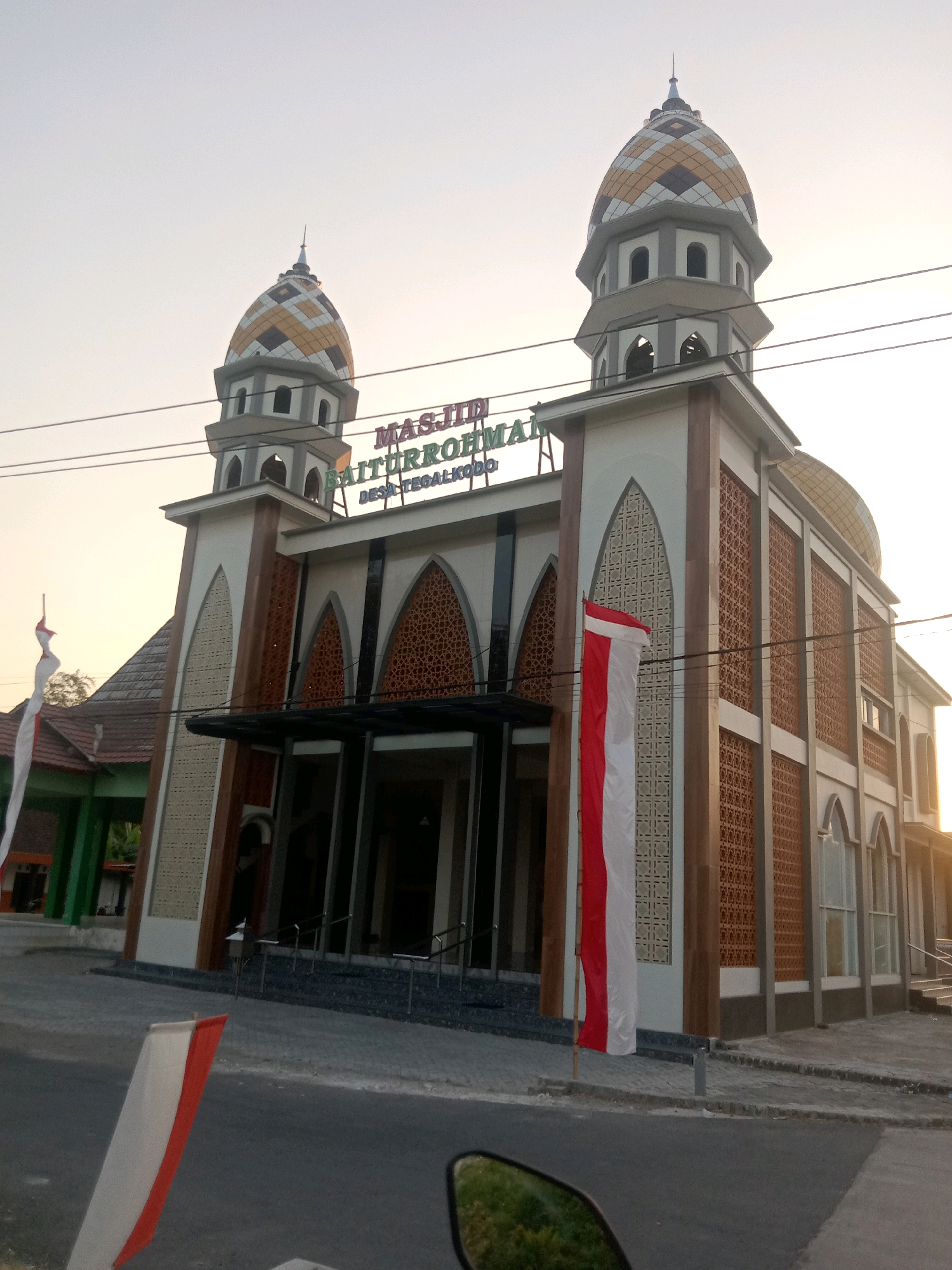 This image captures the exterior of the Masjid Baiturrohmai in Jumput, Indonesia, during what appears to be late afternoon or early evening. The mosque features a symmetrical design with two minarets topped by patterned domes in gold, grey, and white. The central facade has a large entrance area with glass doors and an awning, flanked by tall, arched windows with decorative screens. A prominent sign above the entrance reads "MASJID BAITURROHMAI DESA TEGALKUDO". Two Indonesian flags, red and white, are prominently displayed, one on a flagpole near the entrance and another on the left side of the image, partially obscured. The mosque is situated on a paved area, with a road visible in the foreground. The sky is a pale, hazy blue, suggesting the setting sun. There are no people visible in the immediate vicinity of the mosque, although some structures are visible to the left, suggesting it is part of a village or town. Power lines are also visible across the sky. A portion of a motorcycle's side mirror is visible in the bottom right corner of the frame, indicating the photo was taken from a vehicle.