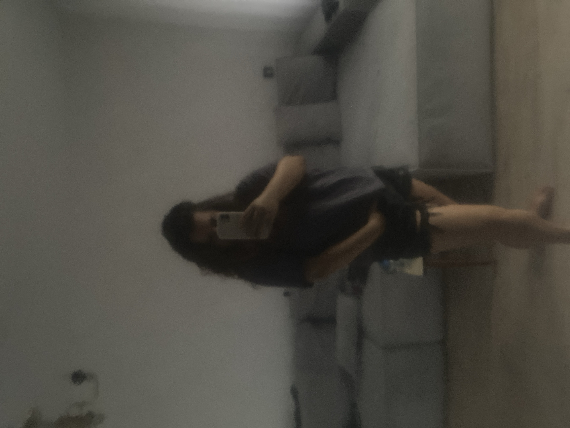 The image is a blurry, full-body mirror selfie taken indoors. A person with long, dark, curly hair is holding a smartphone and appears to be wearing a dark t-shirt and black shorts. The background suggests a living room with a large sectional sofa and a plain wall. The lighting is dim, and the overall image quality is poor, making it difficult to discern fine details. There is no information within the image that can be used to identify the location as the city of Perm, Russia.