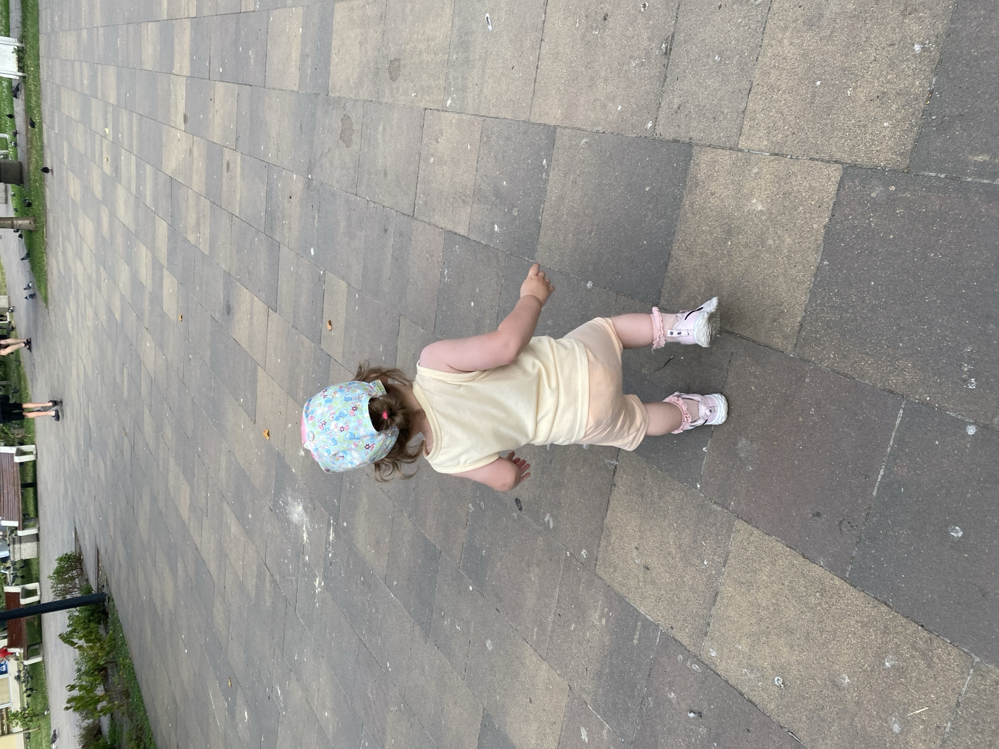 A young child wearing a light yellow sleeveless shirt, light peach shorts, a colorful patterned cap, and pink-ruffled socks with white shoes walks away from the camera on a paved stone pathway in an outdoor setting. The pathway is composed of rectangular gray and tan stones. The child's hair is pulled back in a ponytail. In the background, there are park benches, green bushes, and several pigeons. The lighting suggests it is daytime.