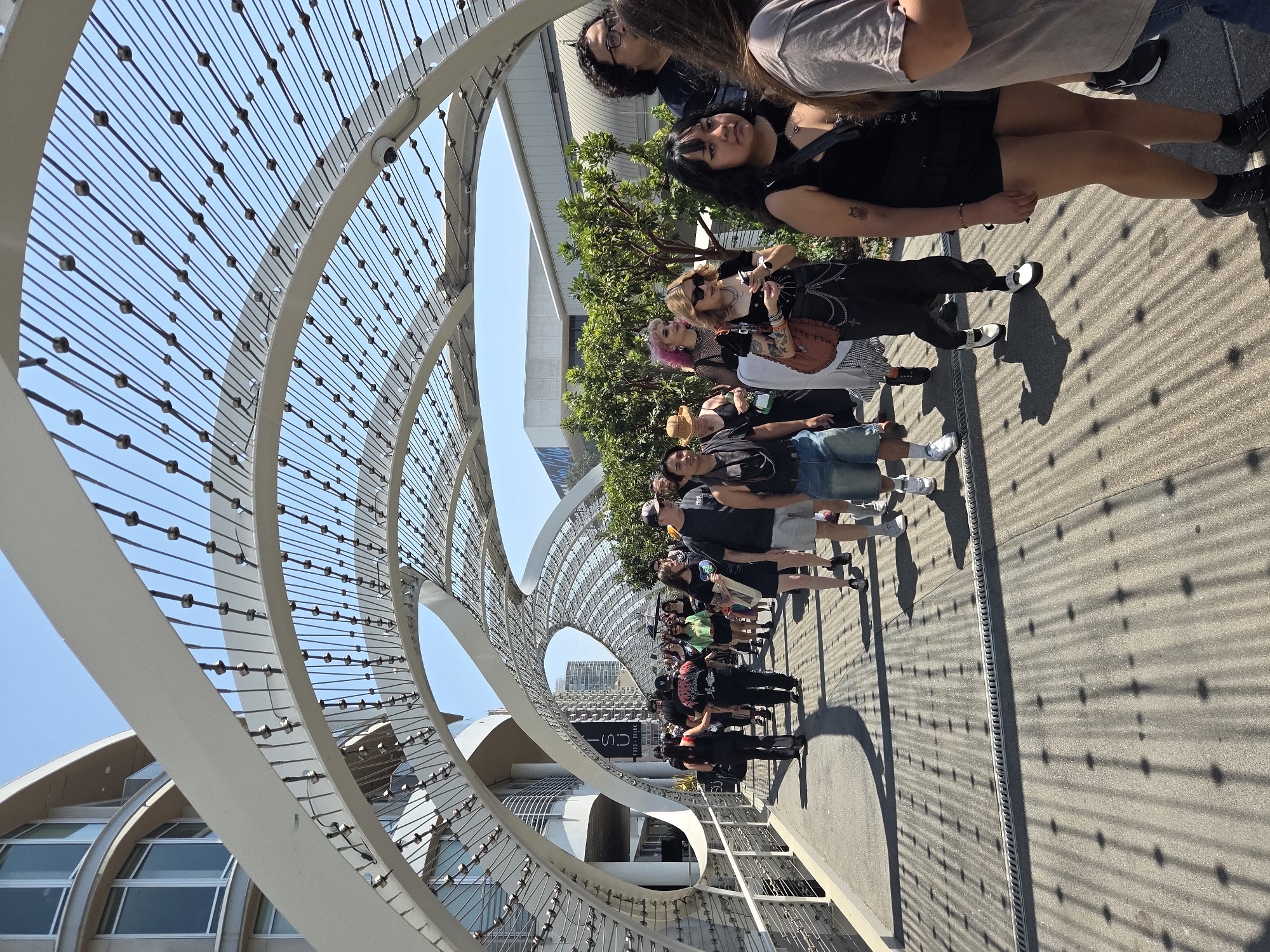 A group of people are walking on a pedestrian bridge in Long Beach, California. The bridge has a white, arched, exoskeleton-like structure overhead with numerous thin cables. The ground is a paved surface with metal grates running horizontally. The people are dressed in casual attire, with some appearing to have a punk or gothic aesthetic. The sun is shining, casting long shadows from the bridge structure onto the ground. In the background, there are buildings, trees, and a clear blue sky. One building in the background has signage that reads "USU FOOD + DRINK". The location is identified as the Long Beach Convention Center.