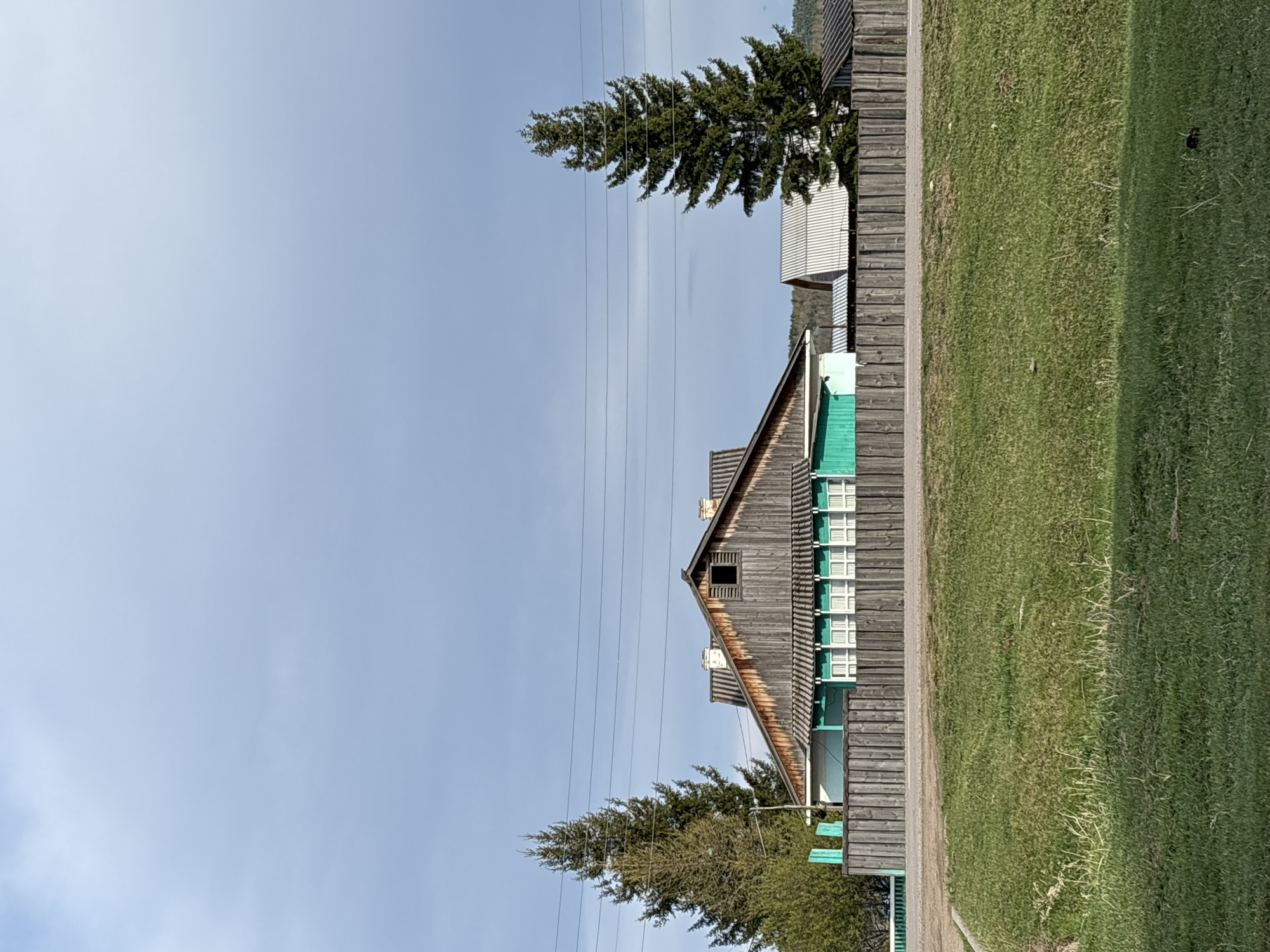 The image depicts a rural scene in Zagatuy, Russia. A wooden building with a distinctive gabled roof and several windows stands alongside a grassy area. The building's wooden exterior has a weathered appearance. The windows are painted with a light green or blue hue, contrasting with the wood. A tall evergreen tree rises to the left of the building, and a smaller structure is partially visible behind it. Overhead power lines cut across the sky, which is a pale blue. The grass is green.