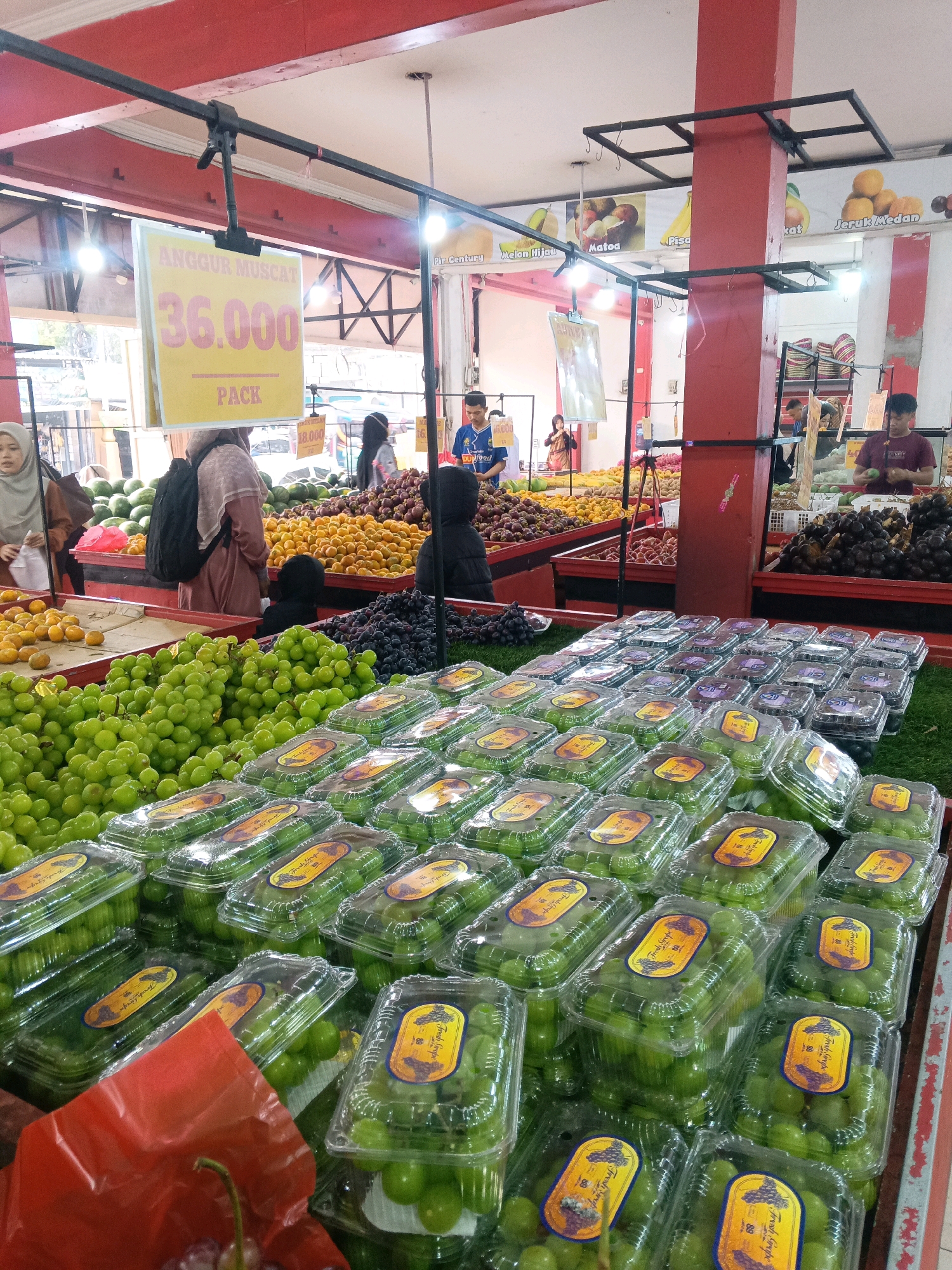 An indoor market scene in Garut, Indonesia, displays a vibrant assortment of fresh fruits. In the foreground, rows of packaged green grapes are meticulously arranged in clear plastic containers, with some loose bunches of grapes visible in the background. To the left, a pile of watermelon and a variety of oranges are showcased. Further back, stalls are laden with other fruits, including dark purple grapes, what appear to be plums or similar dark stone fruits, and baskets of produce. A prominent sign in yellow and pink, hanging from the ceiling, advertises "ANGGUR MUSCAT 36.000 PACK" (Muscat Grapes 36,000 per pack). Several individuals are present, some appearing to be shoppers and others potentially vendors. The environment is brightly lit, suggesting it is daytime. The overall impression is one of a bustling and well-stocked fruit market.