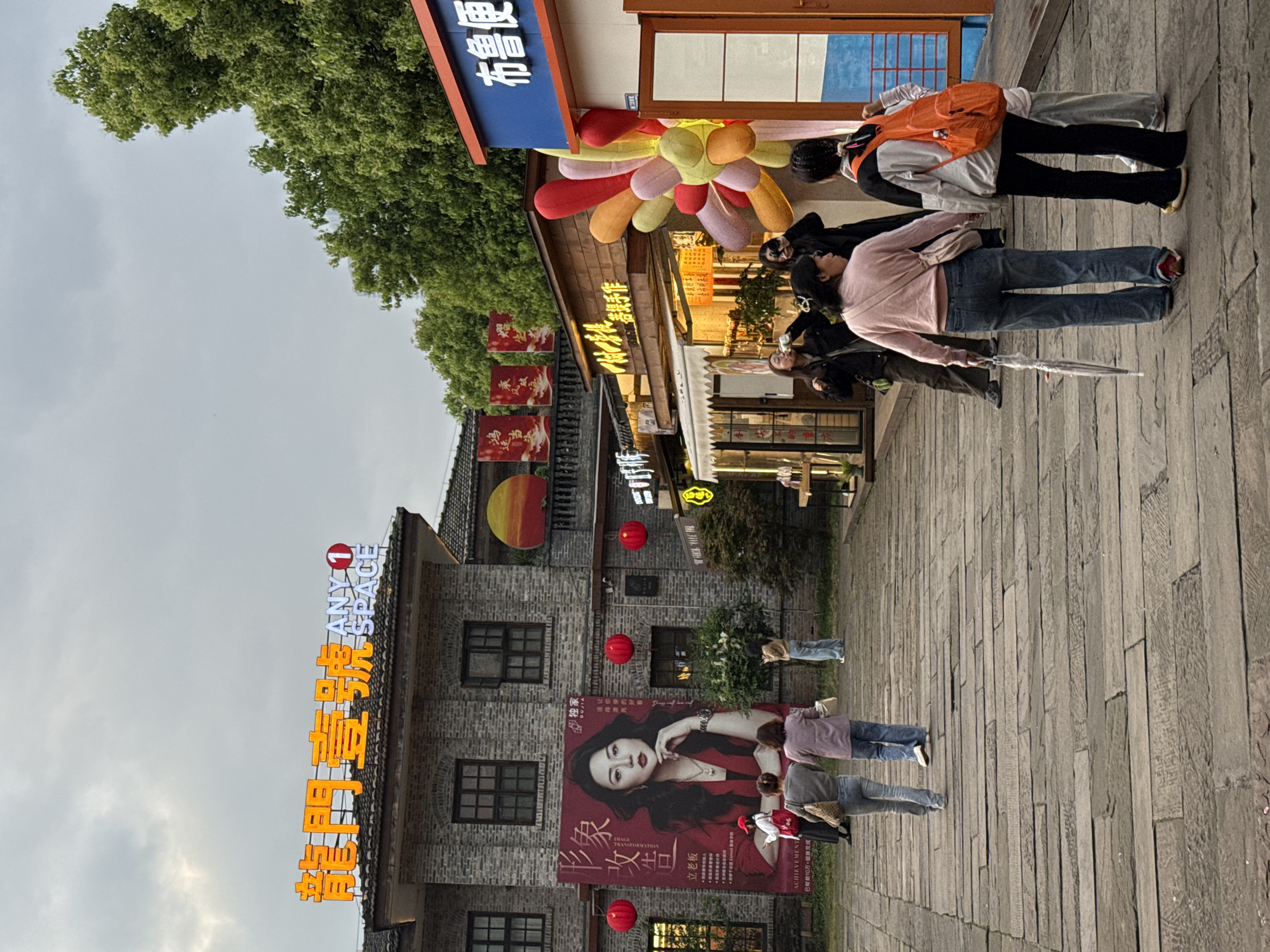 This image depicts an outdoor pedestrian commercial area in Nan'an District, China. The scene features multiple brick buildings with traditional architectural elements under an overcast sky.
On the left, a prominent multi-story brick building displays a large vertical red banner featuring a portrait of a woman with long dark hair, wearing a red top. The banner includes Chinese characters and the English word "ACHIEVEMENT." Three red spherical lanterns are suspended in front of this banner. Above this building, an illuminated sign with orange Chinese characters "龍門壹號" (Longmen Yi Hao) and "ANY SPACE" in white lettering is visible on the rooftop.
To the right, a row of single-story shops with wooden facades and illuminated interiors line the walkway. One shop displays a blue sign with white Chinese characters "布鲁便" (Bu Lu Bian). Adjacent to this shop entrance, a large, colorful inflatable structure resembling a flower, composed of multiple balloon-like petals in shades of red, orange, yellow, and pastel pink/purple, is positioned.
The ground consists of a wide walkway paved with irregular dark stone slabs.
Multiple individuals are present in the scene. In the foreground, with their backs to the viewer, are two women: one wearing a light pink long-sleeved top, blue jeans, and red shoes, carrying a white closed umbrella; the other in a light grey and black jacket, black pants, and an orange backpack with a smaller orange pouch attached. Further back and to the left, two women are walking away, one in blue jeans and a grey top, the other in purple. Near the shops on the right, a small group of people are gathered; one person in a black jacket is holding a camera, another is drinking from a bottle, and a person in light blue pants is observing. Near the large red banner, a person in a red hat is visible, alongside other pedestrians.
The lighting suggests late afternoon or early evening due to the ambient light and illuminated signs. Lush green trees are visible in the background, particularly on the right side.