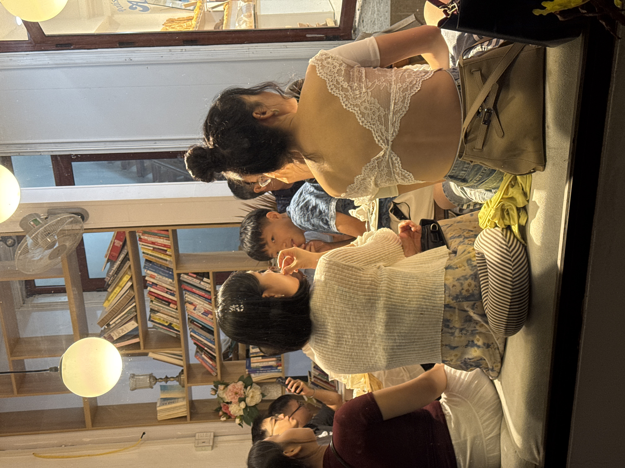 The image shows a group of people seated on a cushioned bench in what appears to be a cafe or restaurant in Mộc Châu, Vietnam. The setting includes bookshelves filled with books, a decorative lamp, and a window overlooking a street. In the foreground, two women are visible from behind, one wearing a white lace top and the other a patterned skirt. Other individuals, including men and women, are seated around a table, engaged in conversation or looking at their phones. The overall atmosphere suggests a casual social gathering.