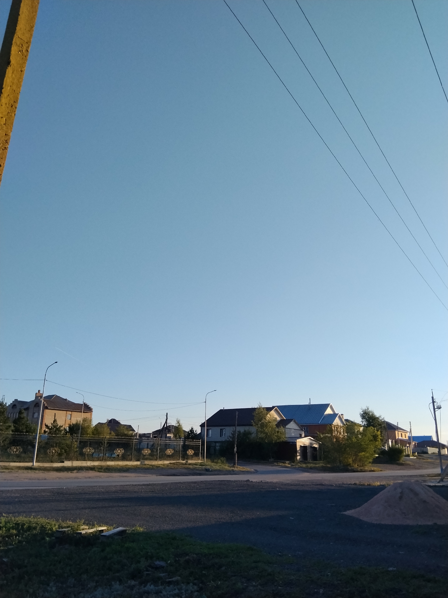 The image captures a street scene in a residential area of Kokshetau, Kazakhstan, under a clear blue sky. On the left, a thick, textured, yellowish pole is partially visible. In the background, a row of houses with various architectural styles and roof types stretches across the frame. Some houses are multi-story, constructed with brick and other materials, and feature windows. A decorative metal fence with ornate scrollwork runs along the front of some properties. Streetlights are present, indicating urban infrastructure. Several trees and bushes are interspersed among the houses. Power lines traverse the upper portion of the image, diagonally crossing the sky. The foreground consists of a dark asphalt road and a strip of grass with some debris. A pile of light-colored gravel is situated on the right side of the foreground. The lighting suggests it is either early morning or late afternoon, with the sun casting gentle shadows. No people are visible in the scene.