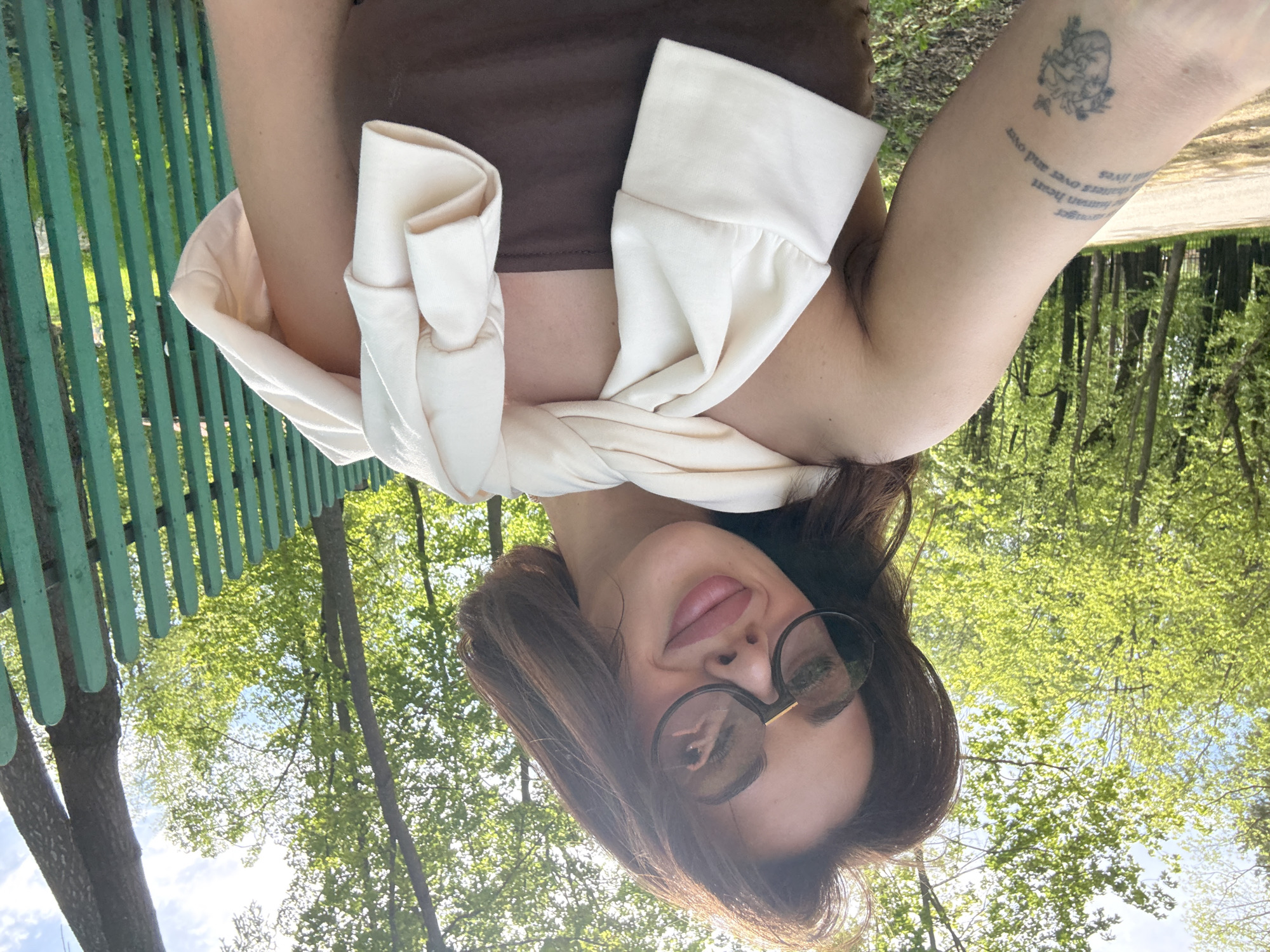A woman is lying on a green park bench outdoors, taking a selfie from below. She is wearing a brown top, with a white sweater wrapped around her shoulders and arms. The woman is wearing glasses. Her left arm, visible in the image, has a tattoo on it. Behind her are green trees and foliage. The location is Sarapul, Russia.